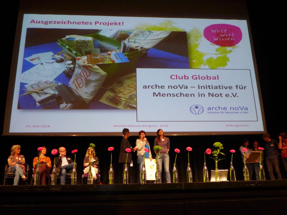 drei Personen stehen auf einer Bühne, hinter ihnen auf der Leinwadn ein Projektion zum Projekt Club Global
