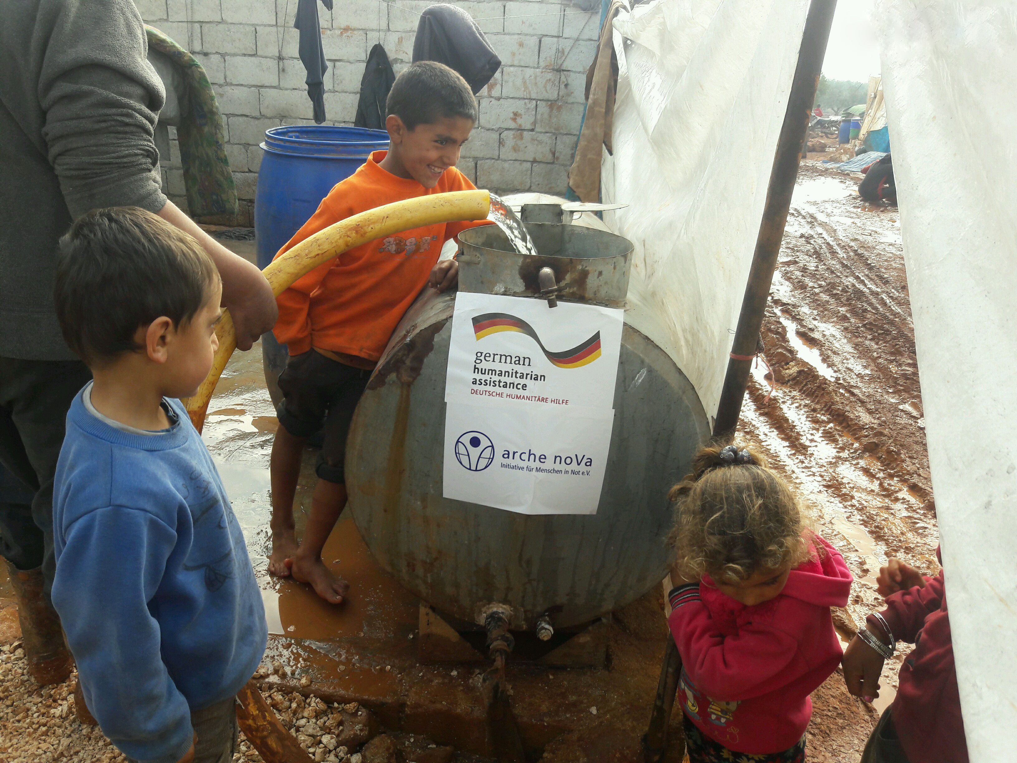 Mann füllt Wasser mit einem Schlauch in einen Tank, barfüßige Kinder stehen im Schlamm daneben