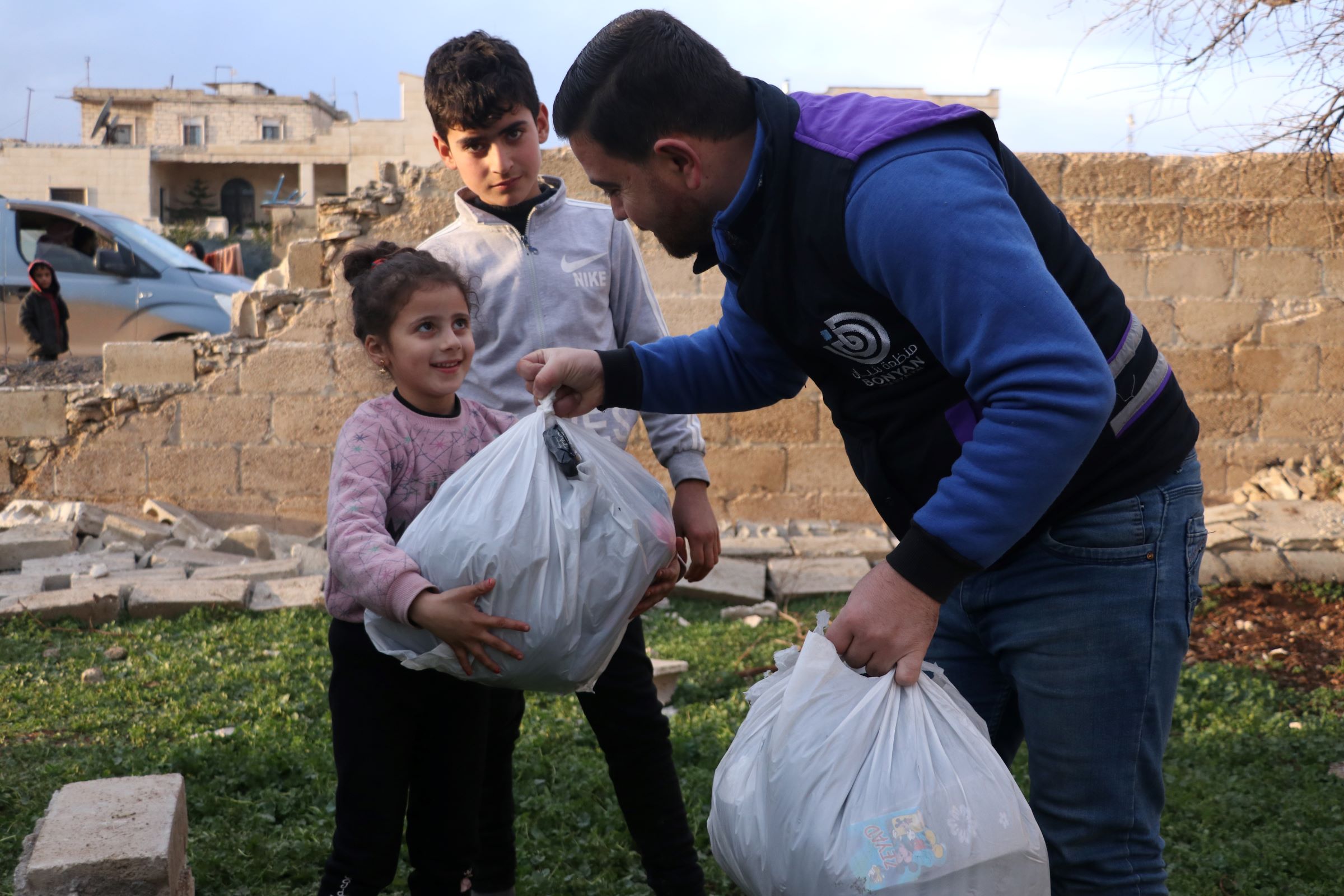 Two children, a girl and an older boy, receive a bag with aid supplies from an emergency response worker of our local partner organization Bonyan. Both are smiling. A collapsed wall can be seen in the background.