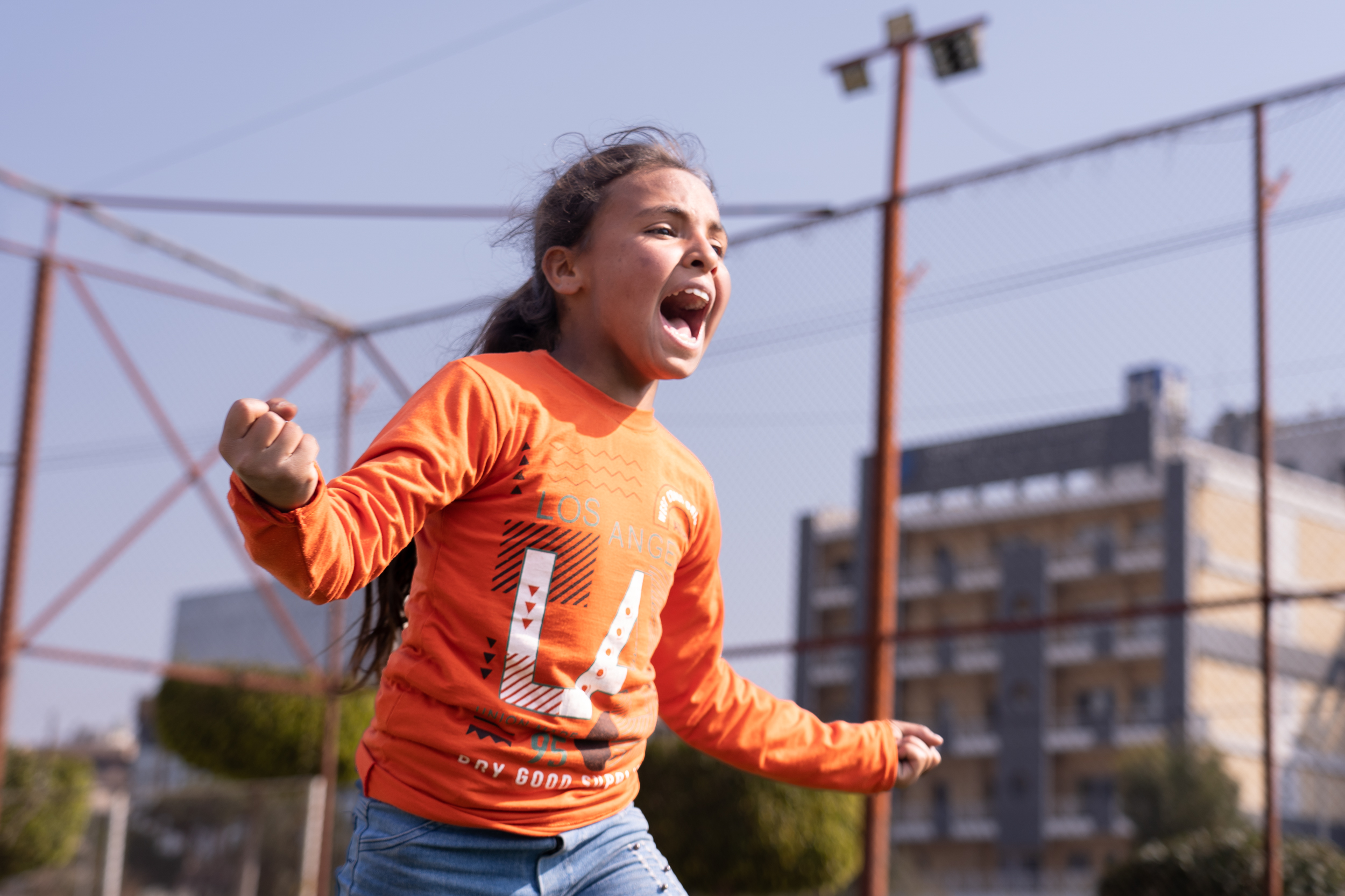 Das Bild zeigt ein Mädchen auf dem Sportplatz, das rennt und gleichzeitig laut ruft. Sie hat beide Arme ausgetreckt und wirkt euphorisch.