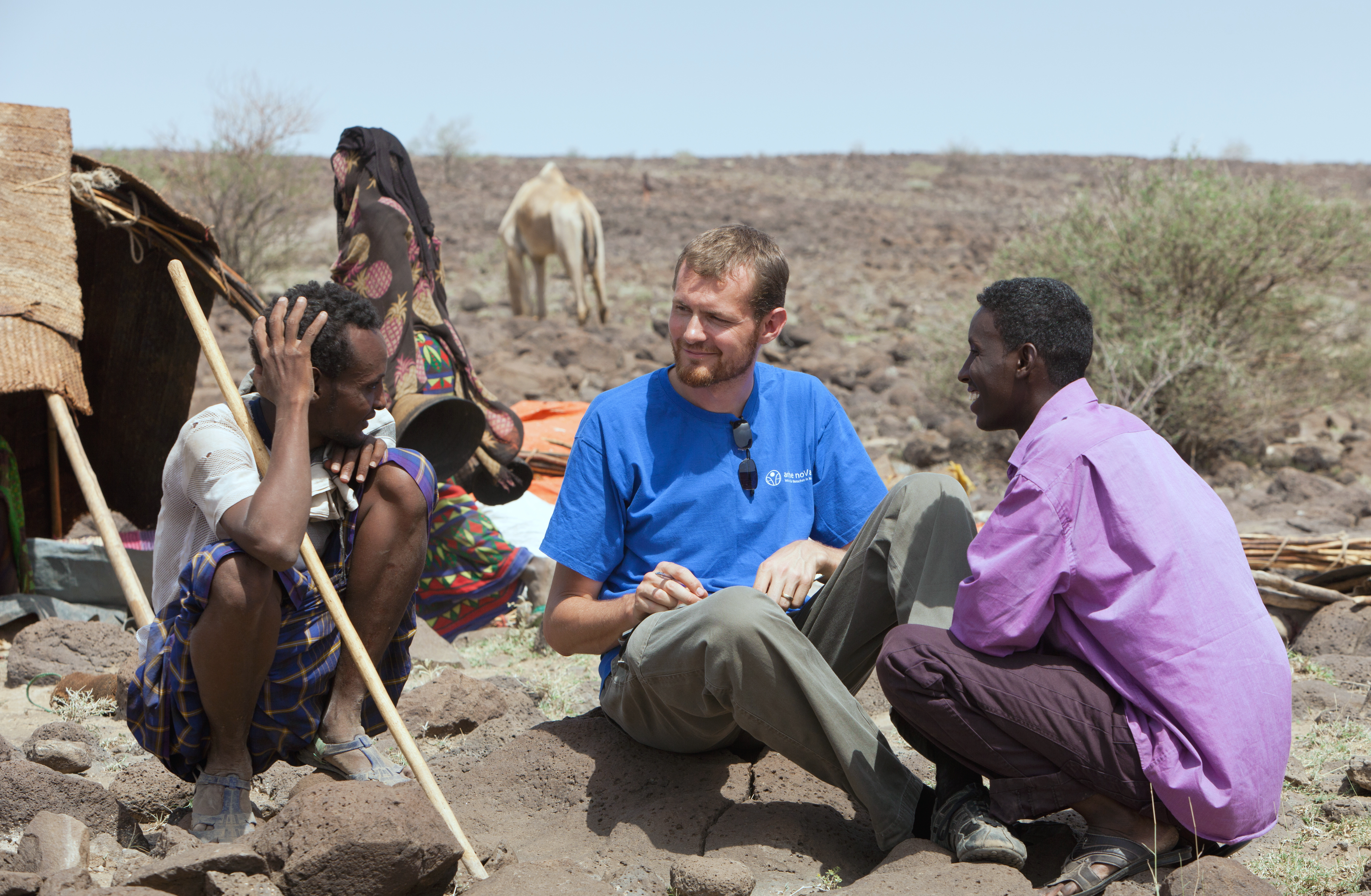 Mathias Anderson (Bild Mitte) sitzt mit zwei anderen Männern auf dem Boden, im Hintergrund Kamele und karge Steppenlandschaft