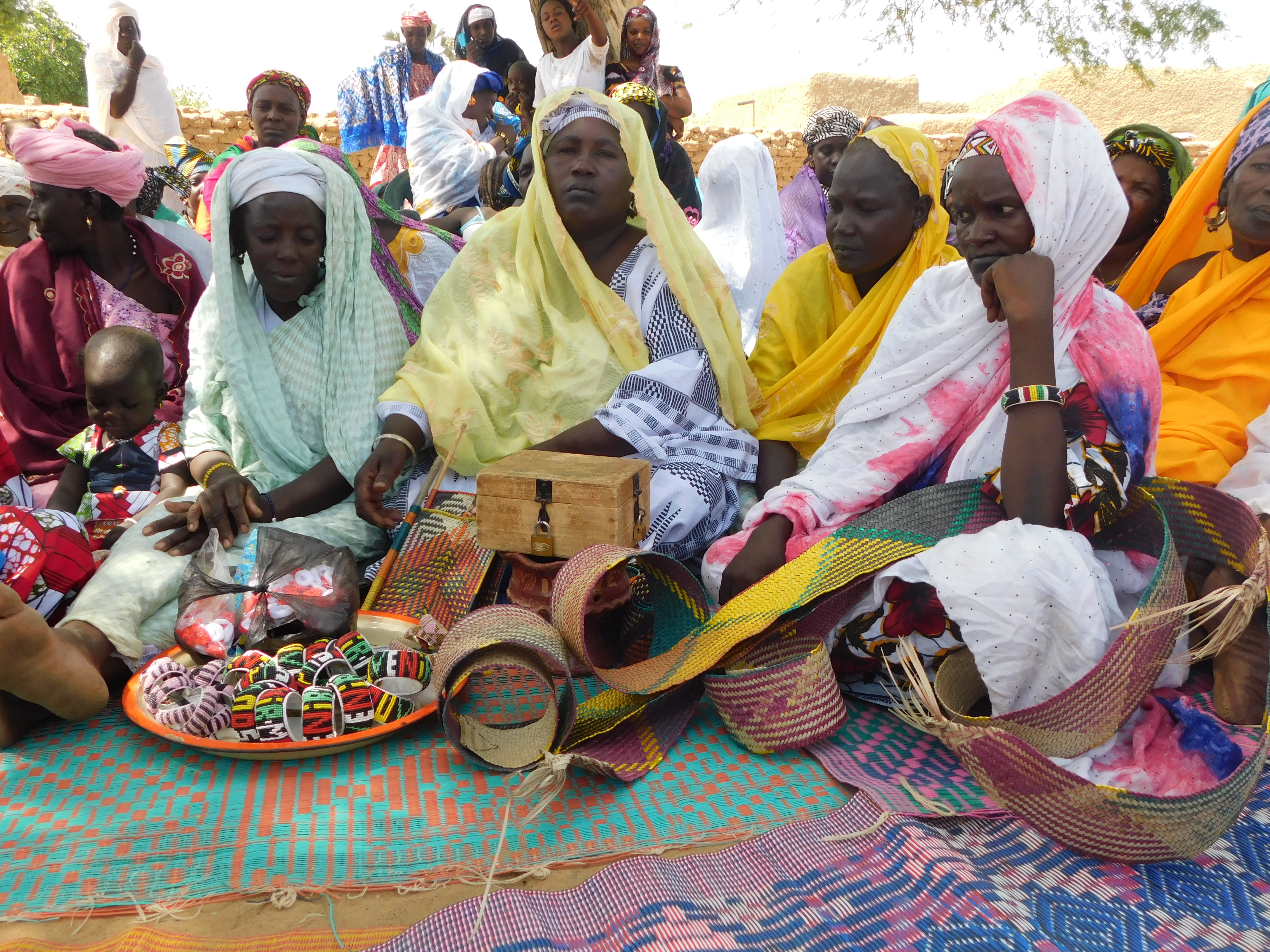 Frauen sitzen auf bunten Matten auf dem Boden. Vor ihnen liegen geflochtene Armbänder und andere kunsthandwerkliche Gegenstände.