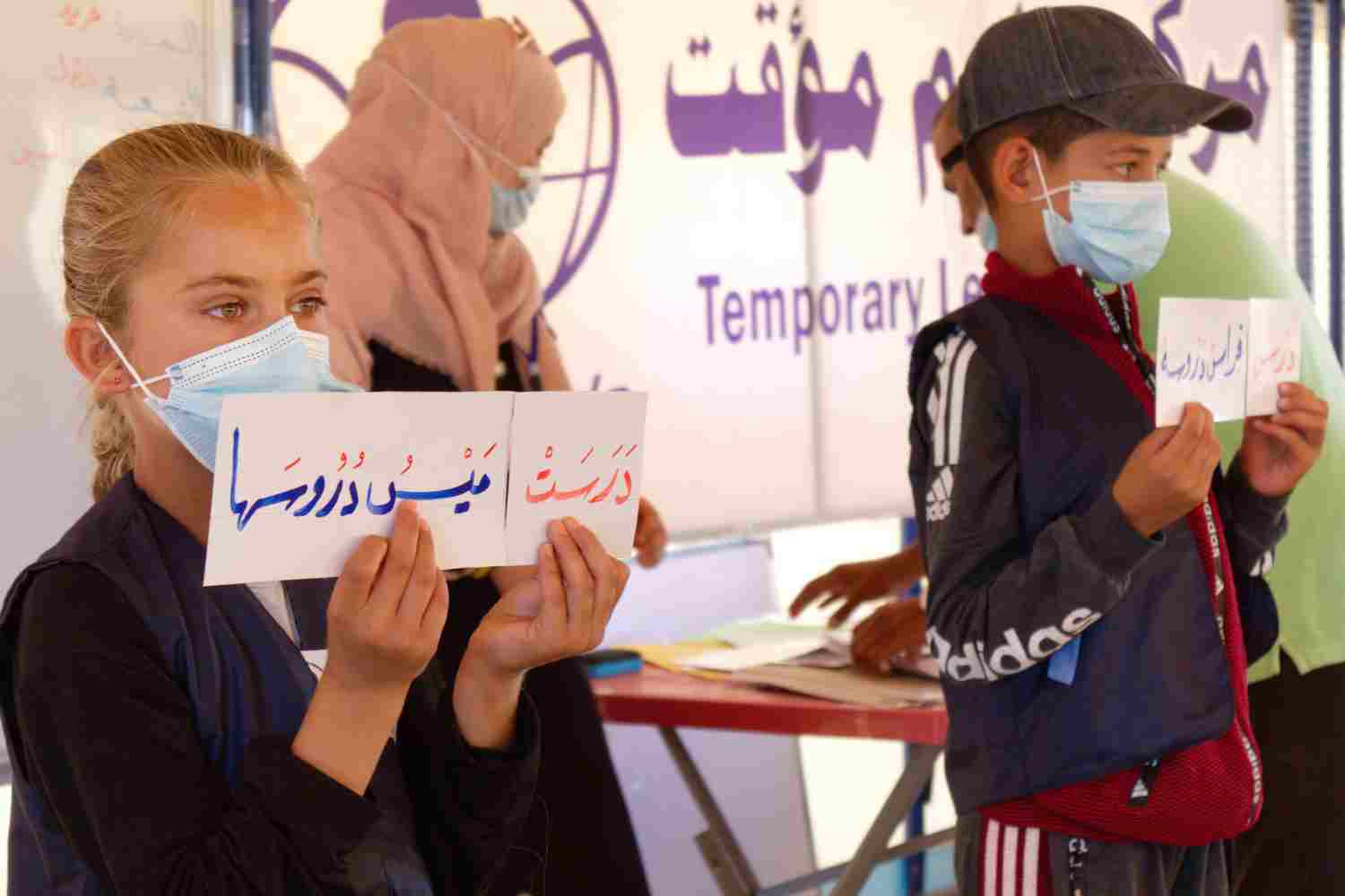 Ein Mädchen hält ein Blatt Papier mit großen arabischen Buchstaben vor sich, weiter hinten ein Junge ebenfalls mit einem Papier