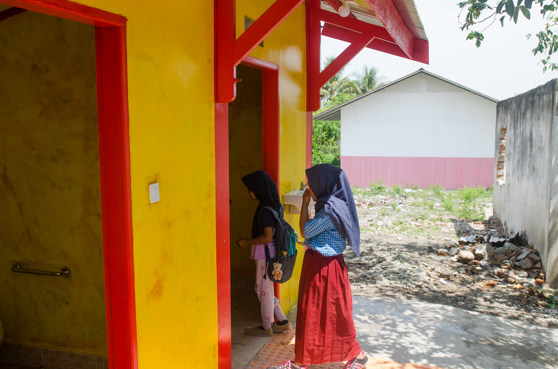 Zwei Mädchen im Schulalter betreten ein gelbes Häuschen durch eine offene Türöffnung.