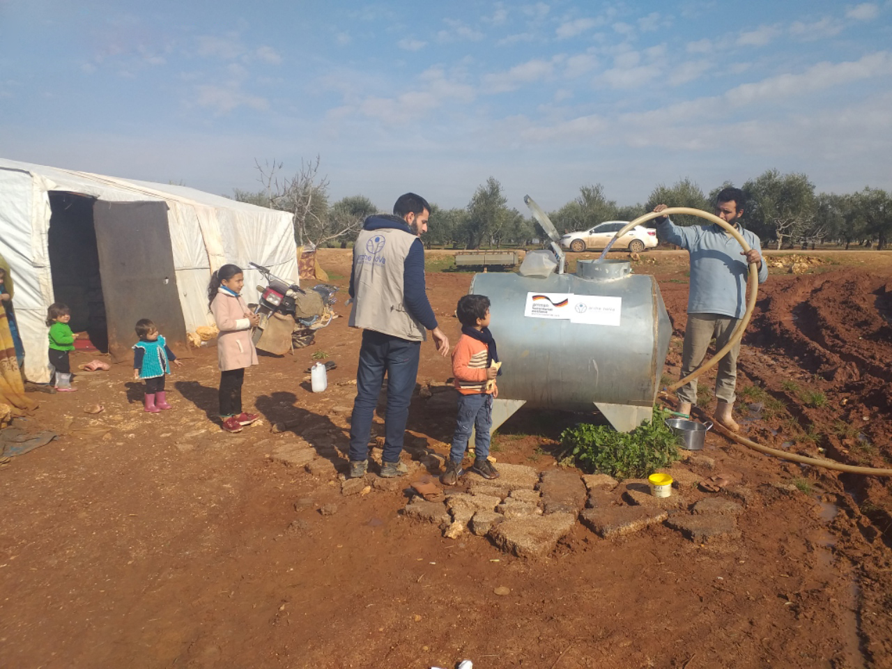 Mann füllt grauen Wassertank mit Schlauch, daneben stehen 4 Kinder und ein Mann und sehen zu