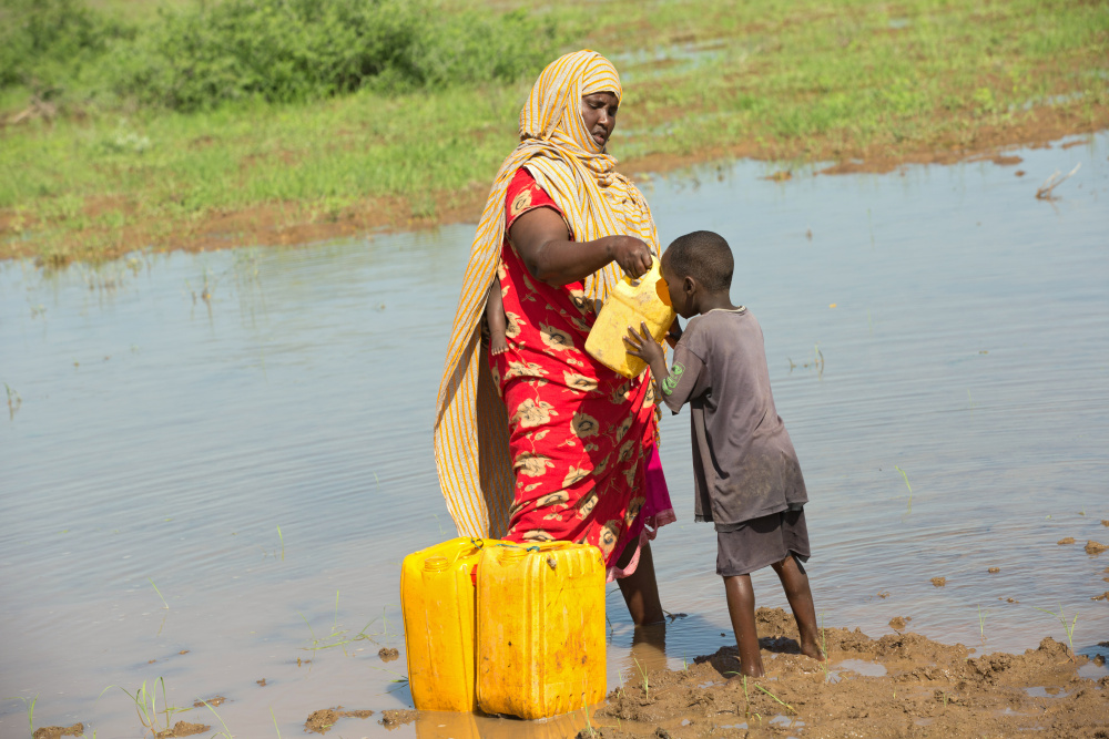 Eine Frau reicht einem Kind einen Behälter zum Trinken, beide stehen mit den Füßen in einem seichten Gewässer