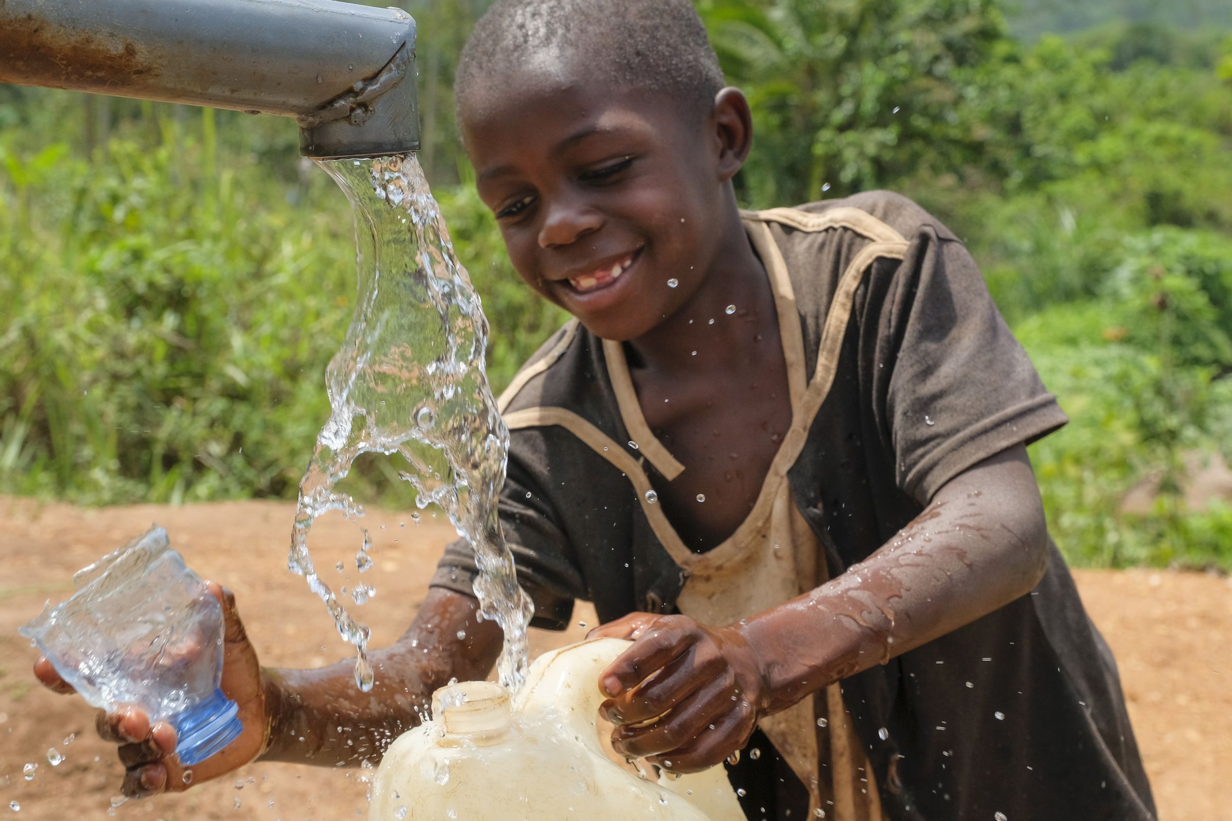 Junge an Wasserhahn einer Pumpe füllt Kanister, Wasserstrahl im Vordergrund