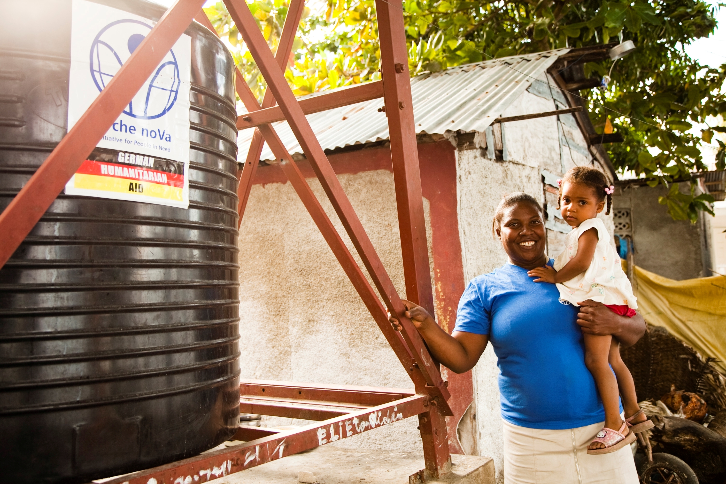 Frau mit Mädchen auf dem Arm lehnt an Stahlkonstruktion, daneben ein Wassertank mit arche noVa Logo