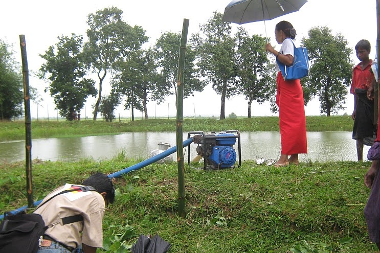 Frau mit Regenschirm an Wasserteich, daneben eine Pumpe mit Schlauch