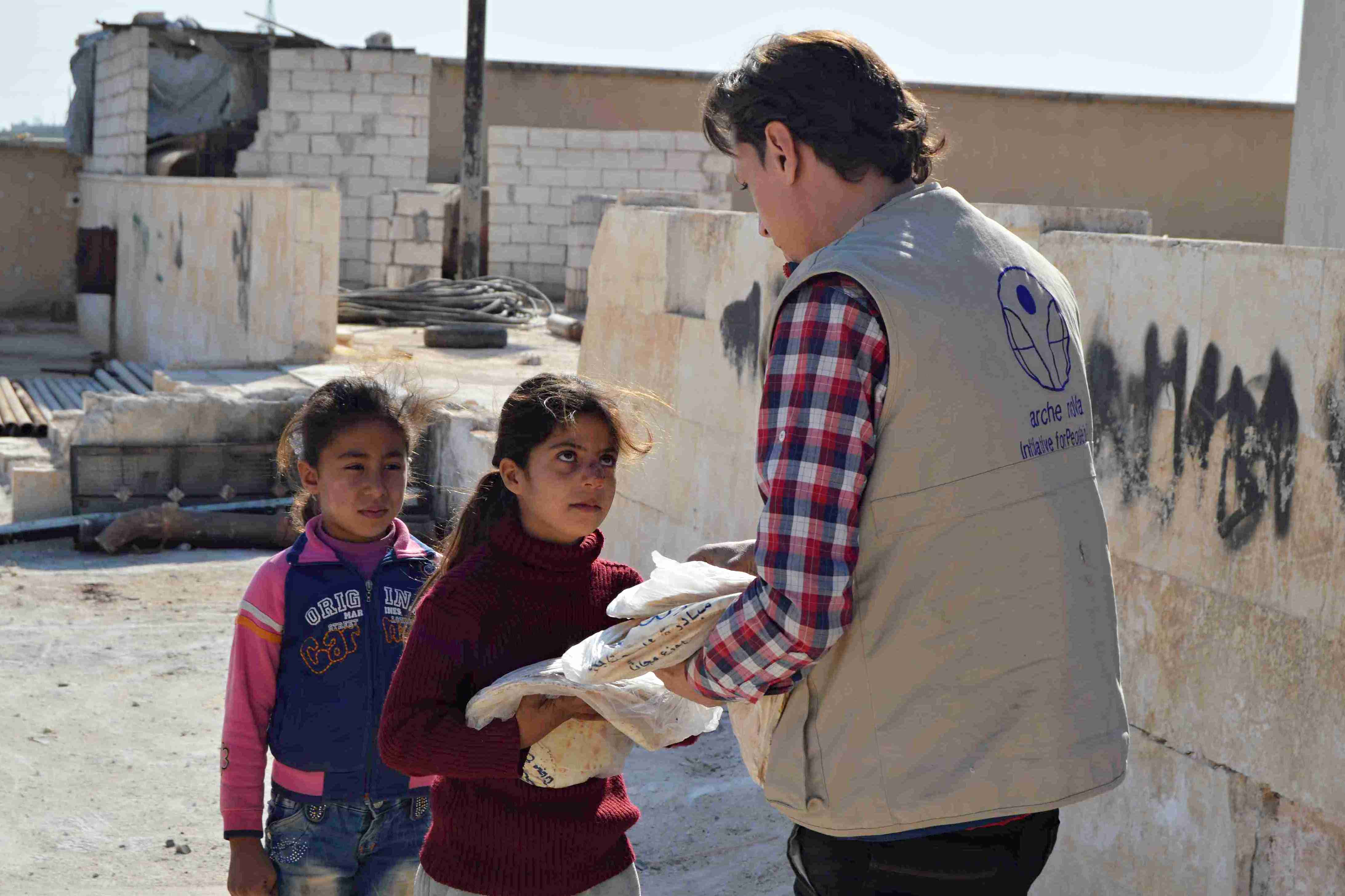 Ein Mitarbeiter des syrischen arche noVa Teams reicht einem Mädchen ein Brotpaket im Hintergrund eine Buruine