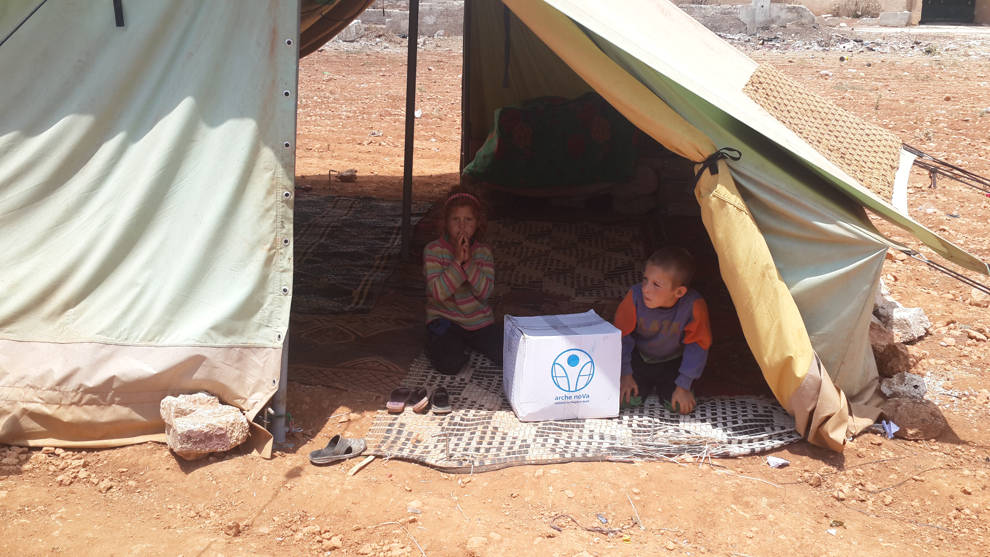 Zwei Kinder sitzen in einem offenen Zelt, zwischen ihnen liegt ein Paket mit Hilfsgütern von arche noVa