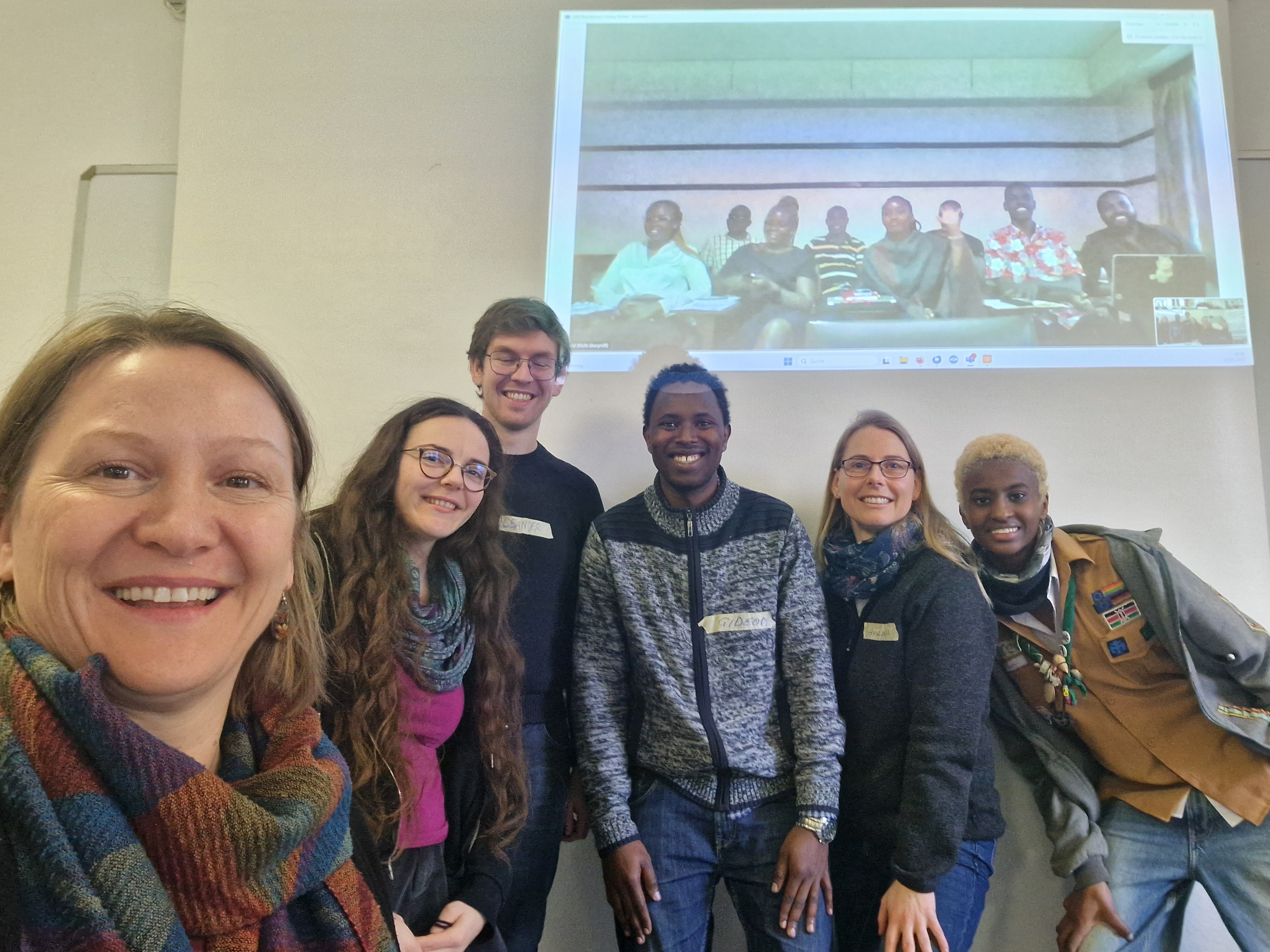 Selfie einer Gruppe von 6 Personen. Im Hintergrund ist die Aufnahme einer weiteren Gruppe an die Wand projiziert.