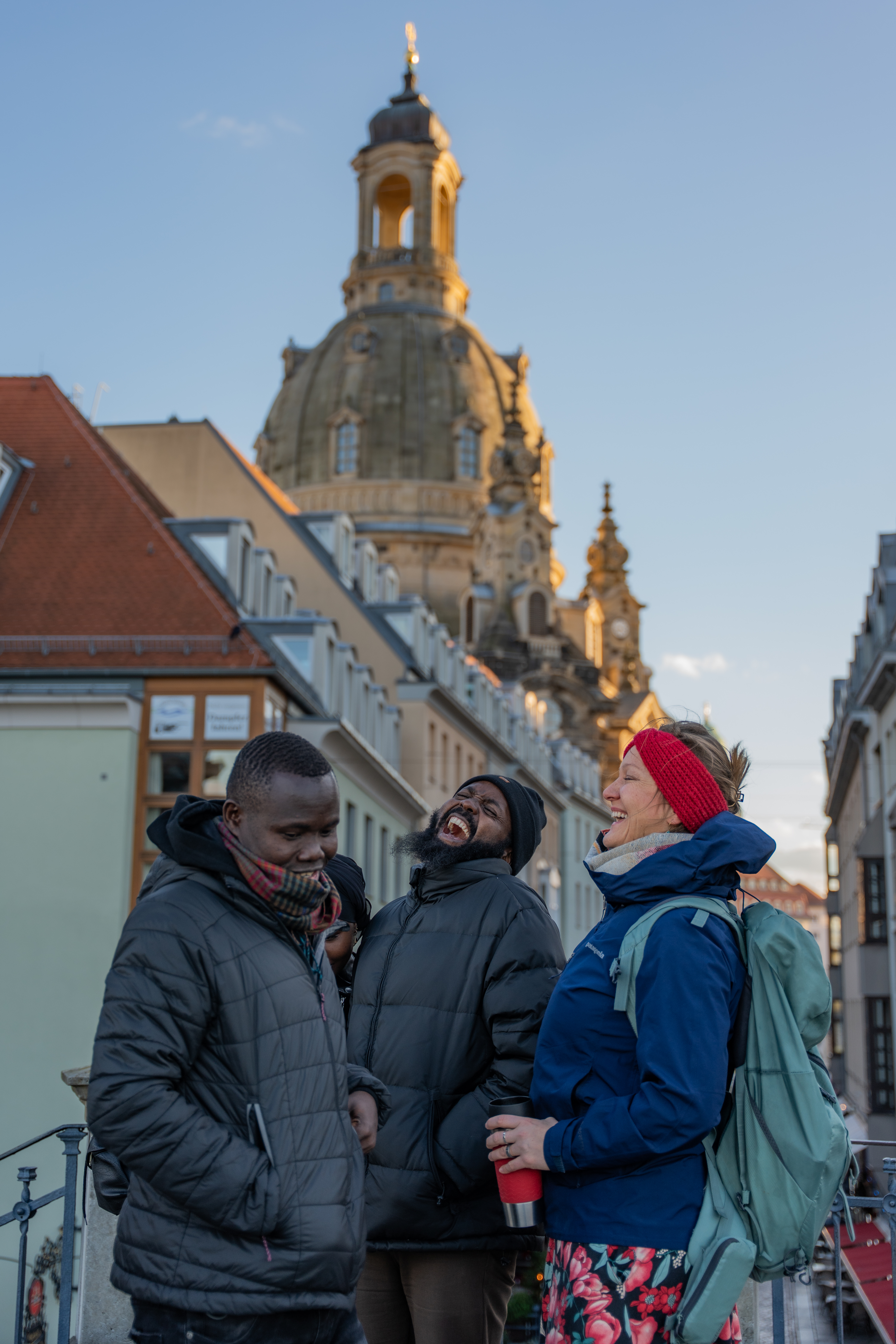 Drei Personen stehen zusammen und lachen. Im Hintergrund ist die Dresdner Frauenkirche zu sehen.