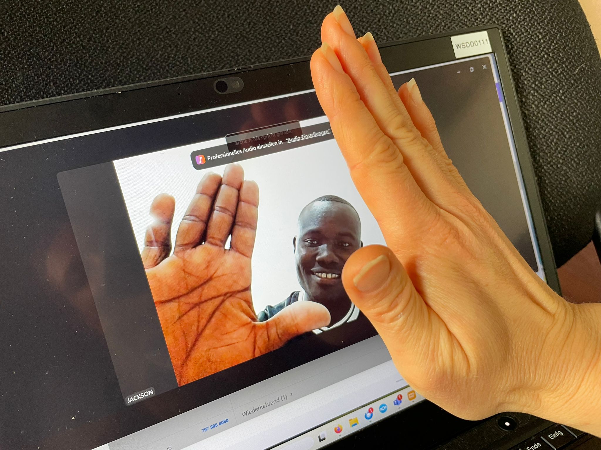 Eine Hand deutet eine High-Five-Geste in Richtung eines Monitors an. Auf dem Monitor ist eine Person zu sehen, die diese Geste ebenfalls andeutet.