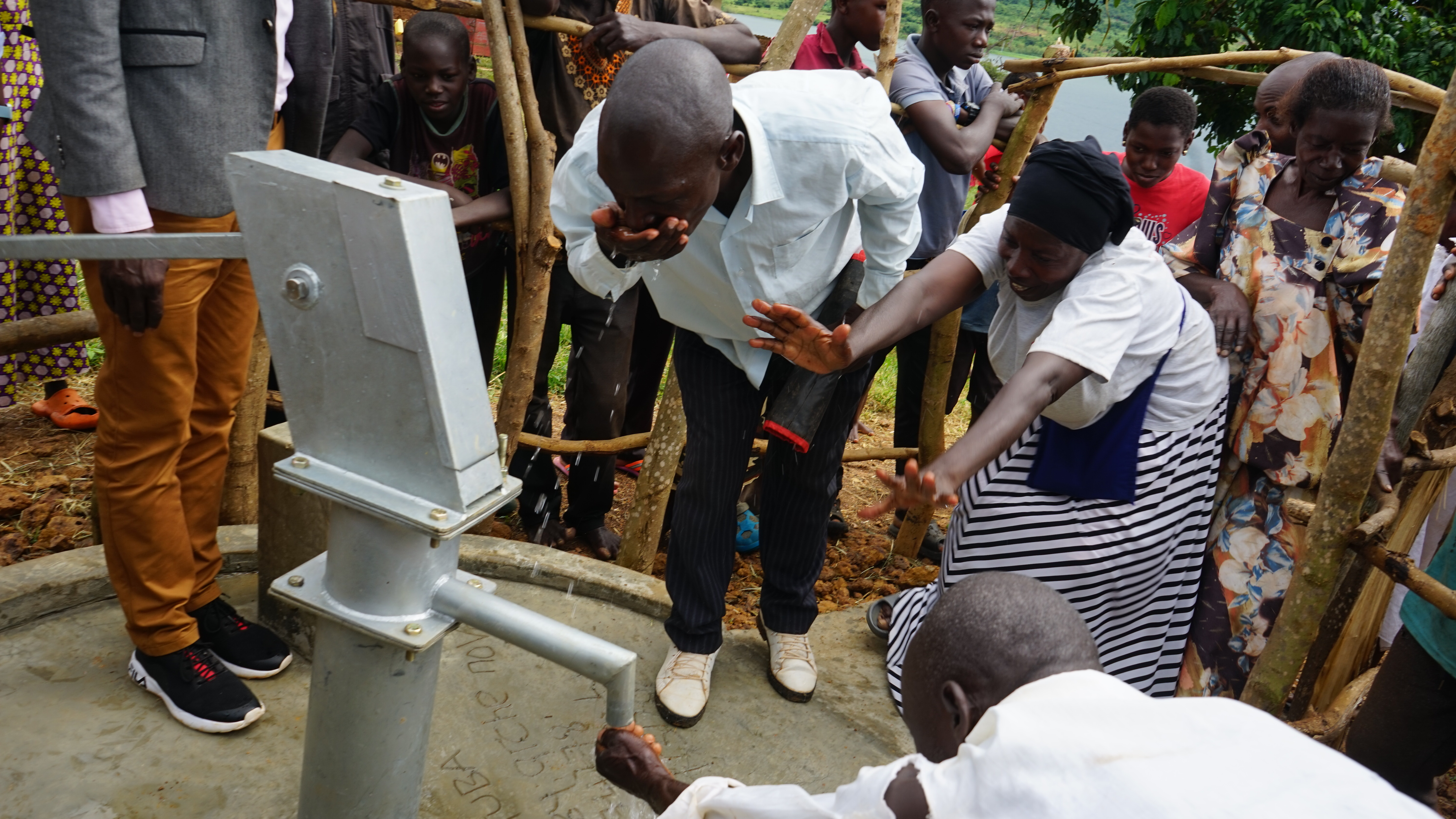 Menschen an einer Handpumpe, ein Mann trinkt aus seiner Handfläche, eine Frau streckt die Arme nach der Pumpe aus