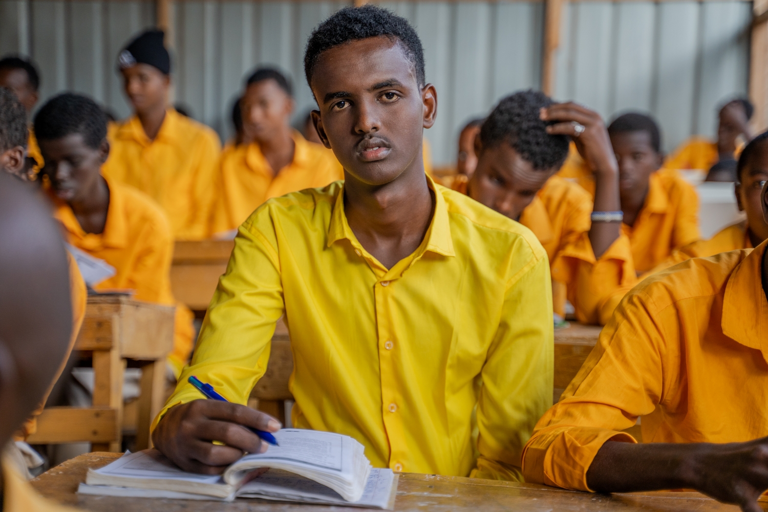 Portrait von Abdifatah an seiner Schulbank. Im Hintergrund sieht man weitere Schüler. 