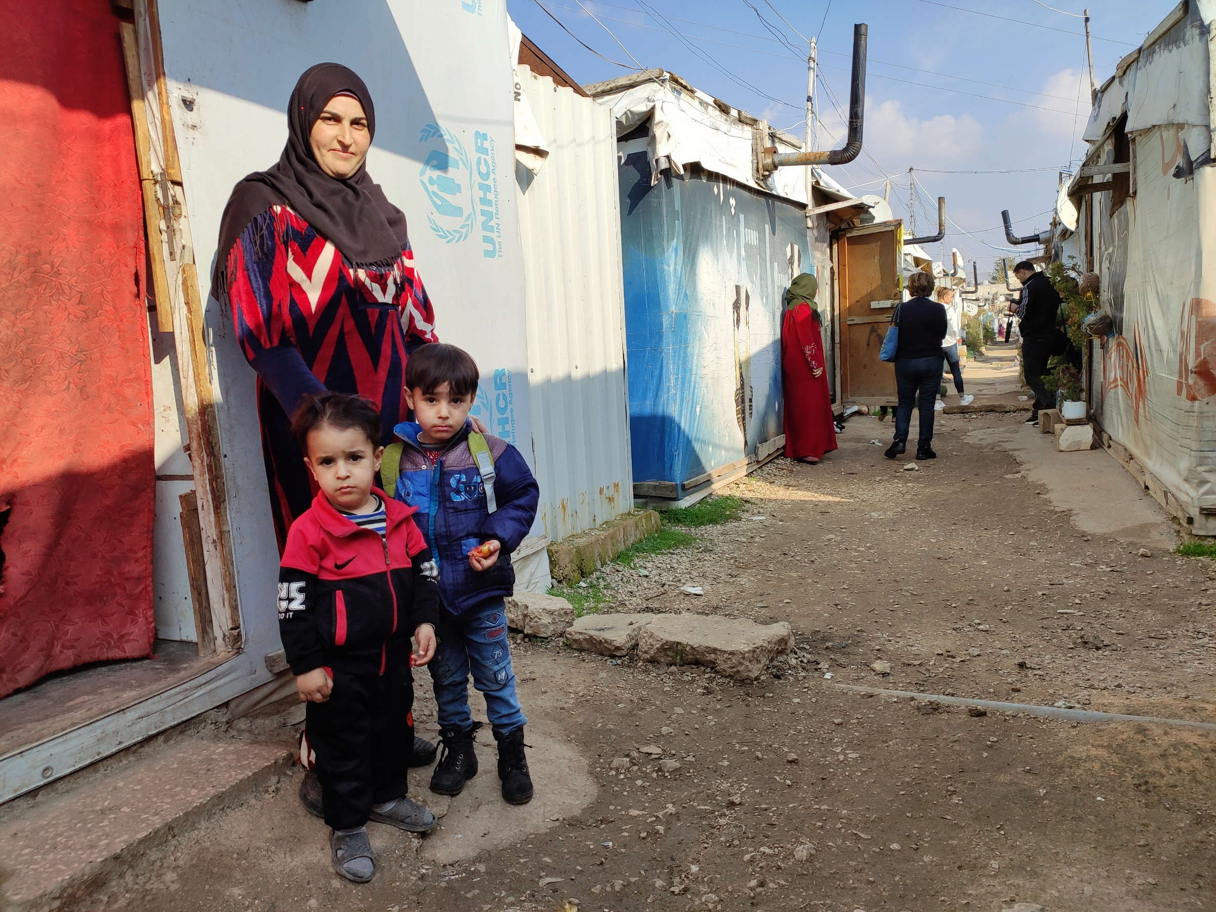 Zwei Kinder vor einer Frau, die vor Behausungen aus Wellblech steht, im Hintergrund eine weitere Menschengruppe
