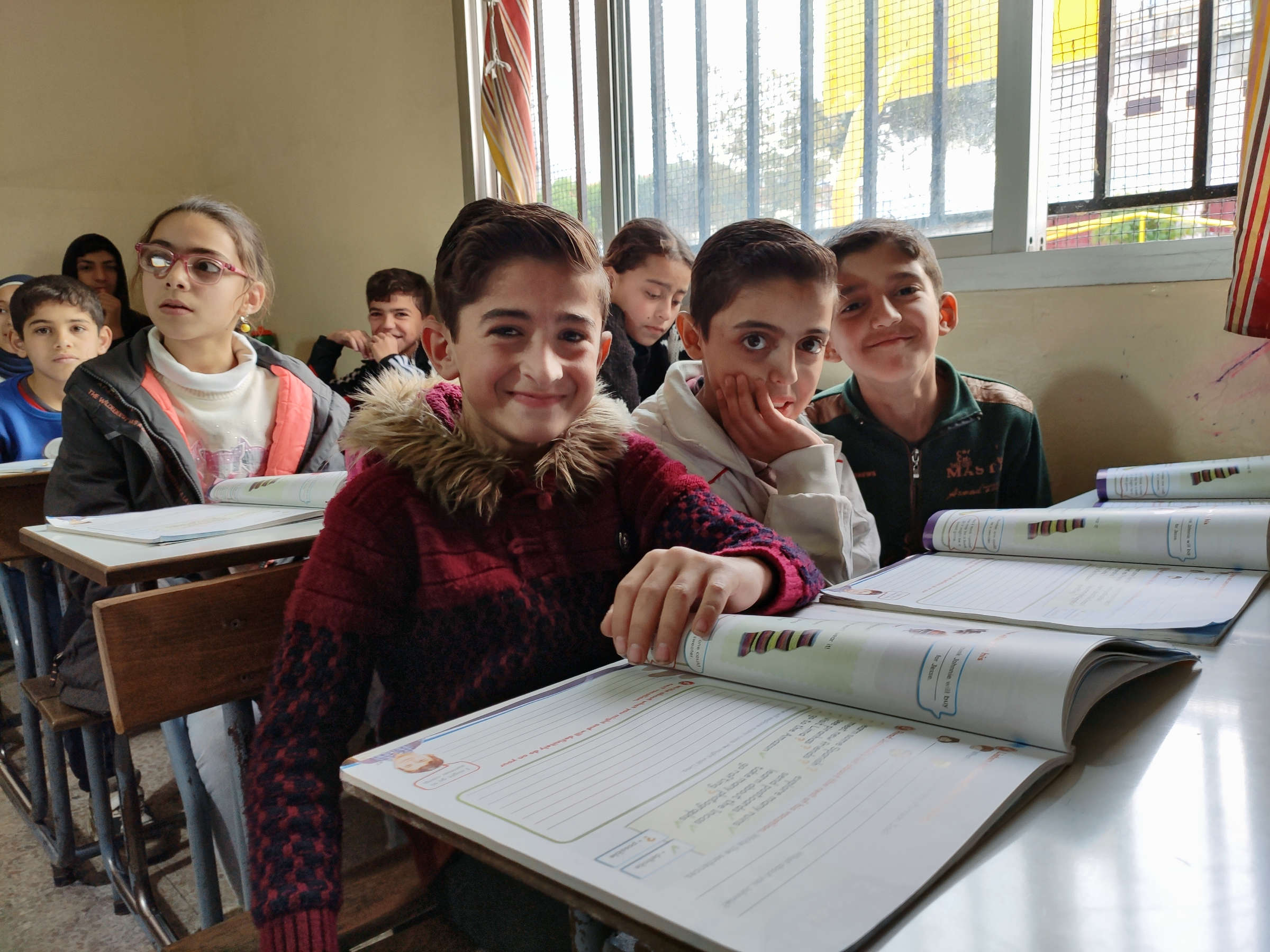 Schülerinnen und Schüler in Klassenraum. Drei Kinder im Vordergrund blicken lächelnd in die Kamera