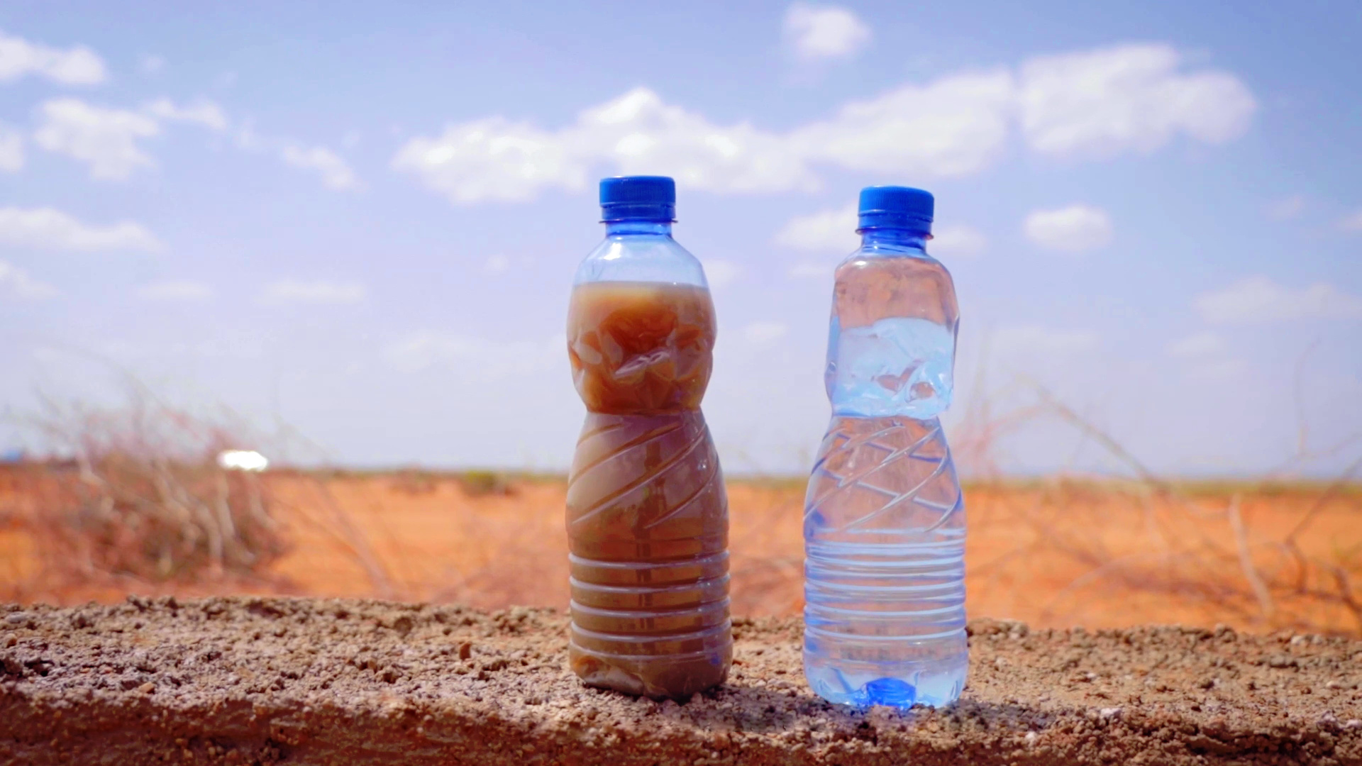 Zwei Kunststoffflaschen auf Sandboden vor blauem Himmel. Die rechte Flasche gefüllt mit undurchsichtiger, brauner Flüssigkeit, die linke mit glasklarer Flüssigkeit