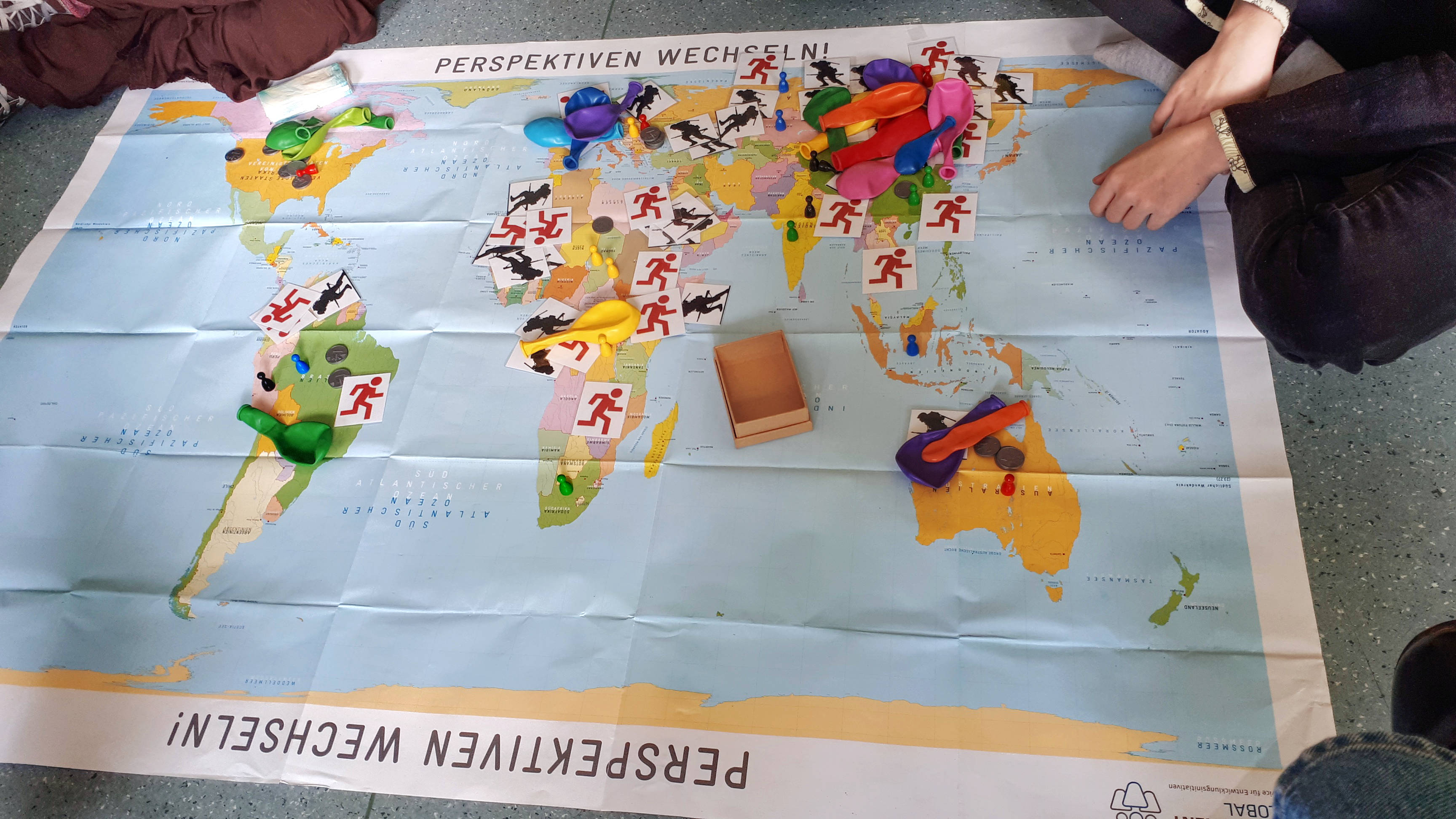 Zentral im Bild liegt eine Weltkarte auf dem Boden, auf der unterschiedliche Dinge auf den einzelnen Kontinenten liegen. Es sind Luftballons, Abbildungen von Soldaten und Geldmünzen zu sehen.