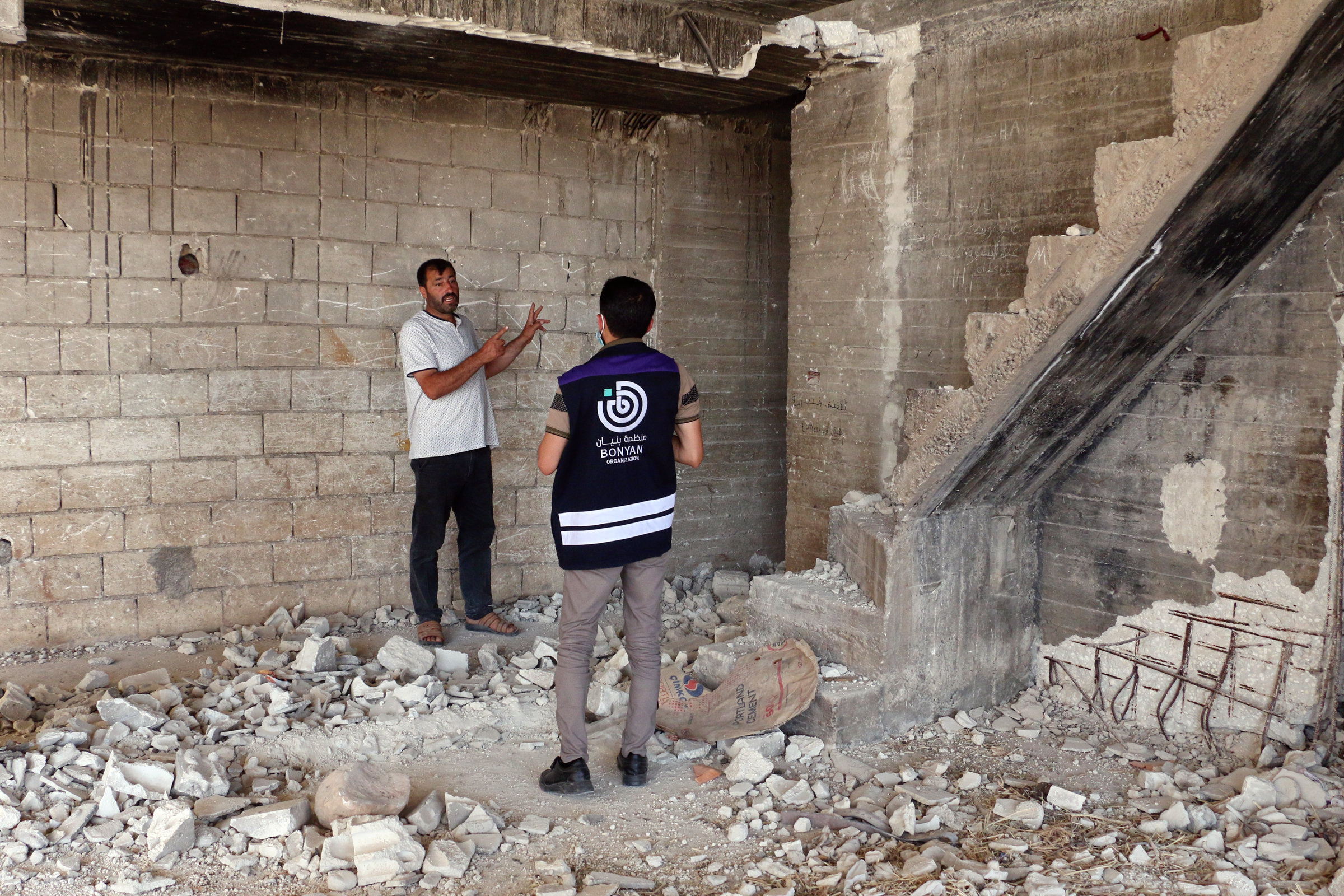 Ahmed Hassan und ein Mitarbeiter von Bonyan stehen in einem offensichtlich zerstörten Haus. Am Boden liegen Trümmer, rechts sieht man eine stark beschädigte Treppe. Hinter Ahmed ist eine offene Ziegelwand zu sehen.