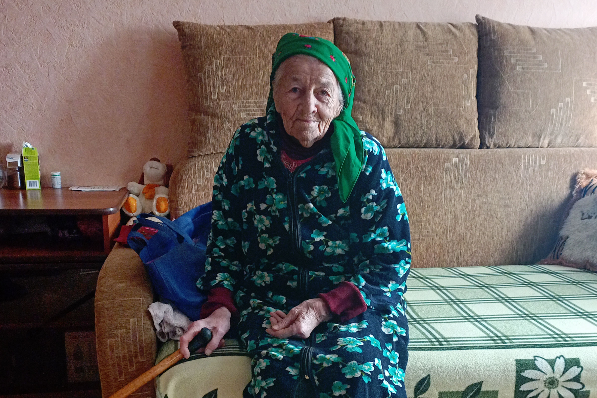 •	Eine ältere Frau sitzt auf einem Sofa. Sie hat einen Gehstock in der Hand. 