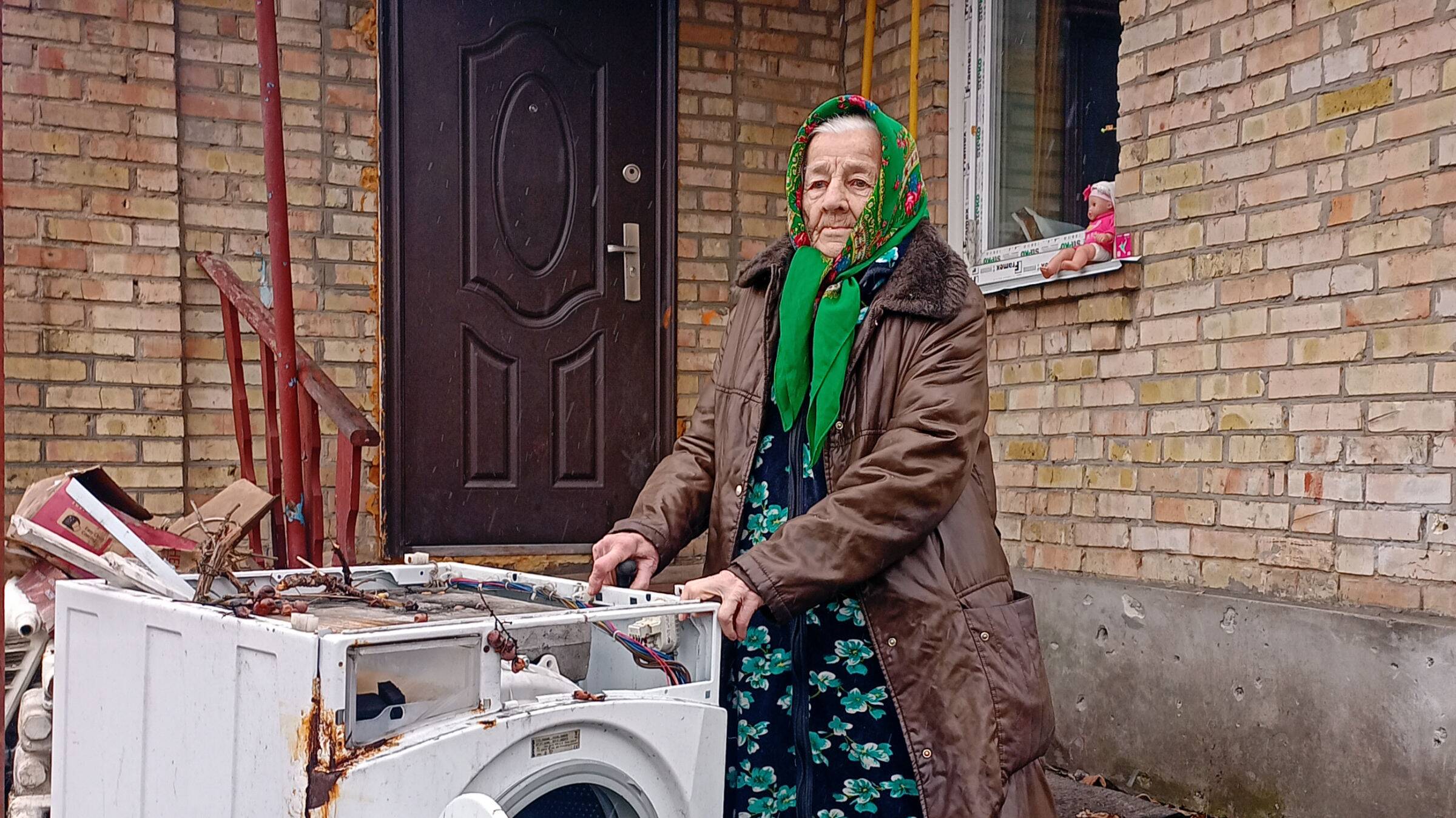Ältere Frau mit einem Kopftuch steht vor einem Haus. Neben ihr ist eine Waschmaschine, die völlig zerstört ist. Die Frau berührt die Waschmaschine mit den Händen.