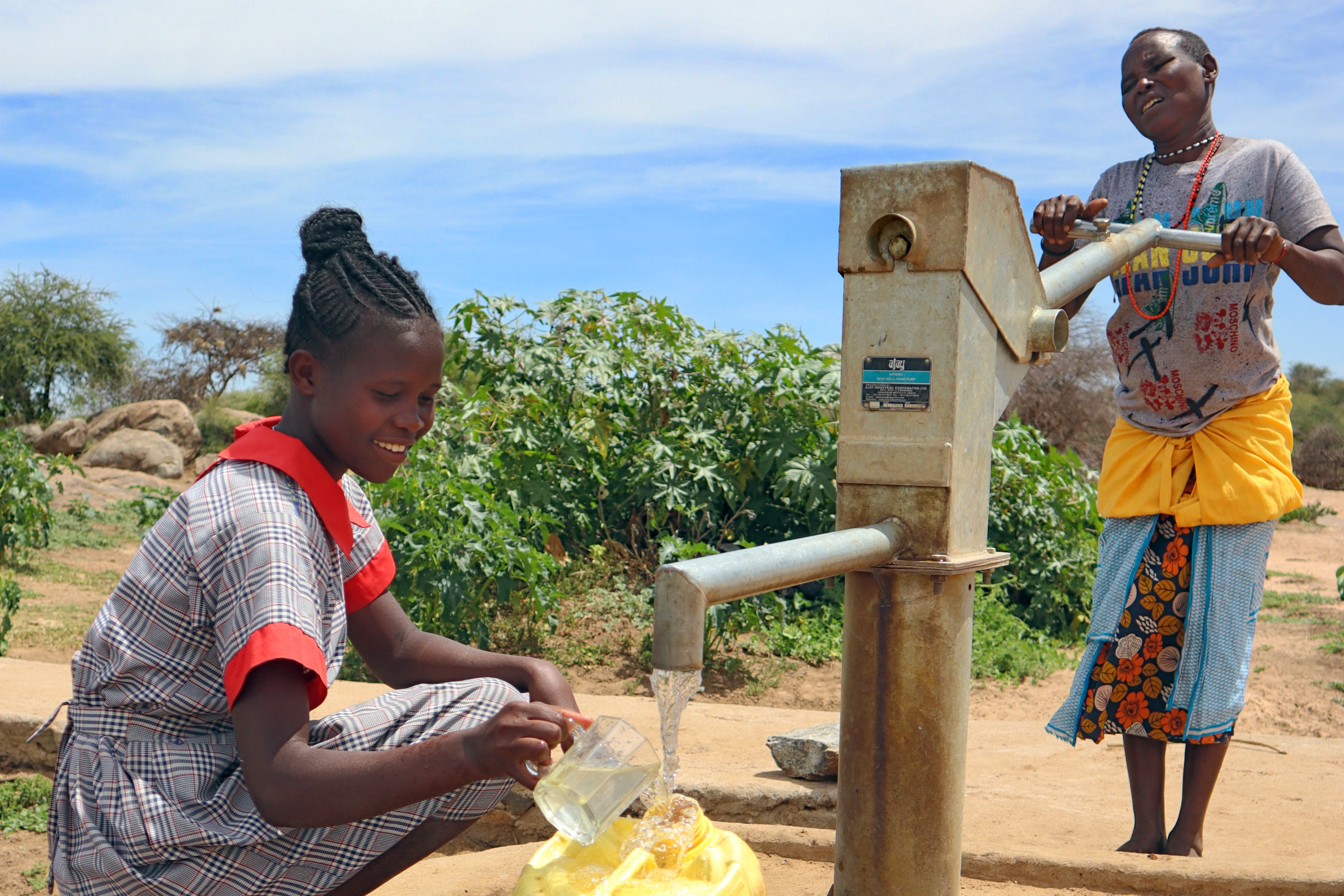 Zwei junge Frauen an einer Handpumpe, eine im Vordergrund hockt beim Hahn, wo Wasser in einen gelben Kanister strömt, die andere hält den Schwengel der Pumpe in beiden Händen