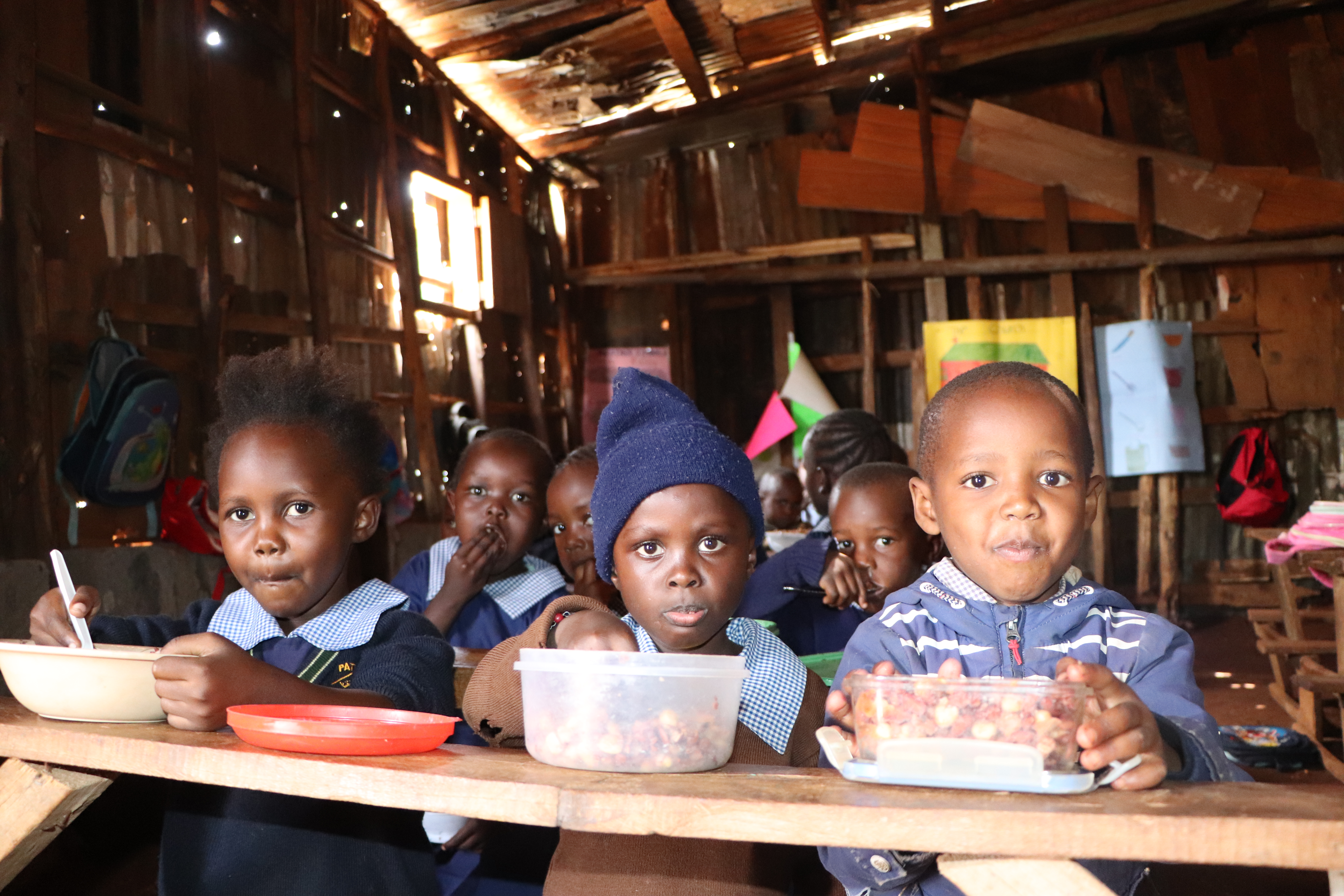 Drei Grundschulkinder sitzen an einer Schulbank und blicken in die Kamera. Auf dem Tisch davor stehen Kunststoffbehälter mit Essen