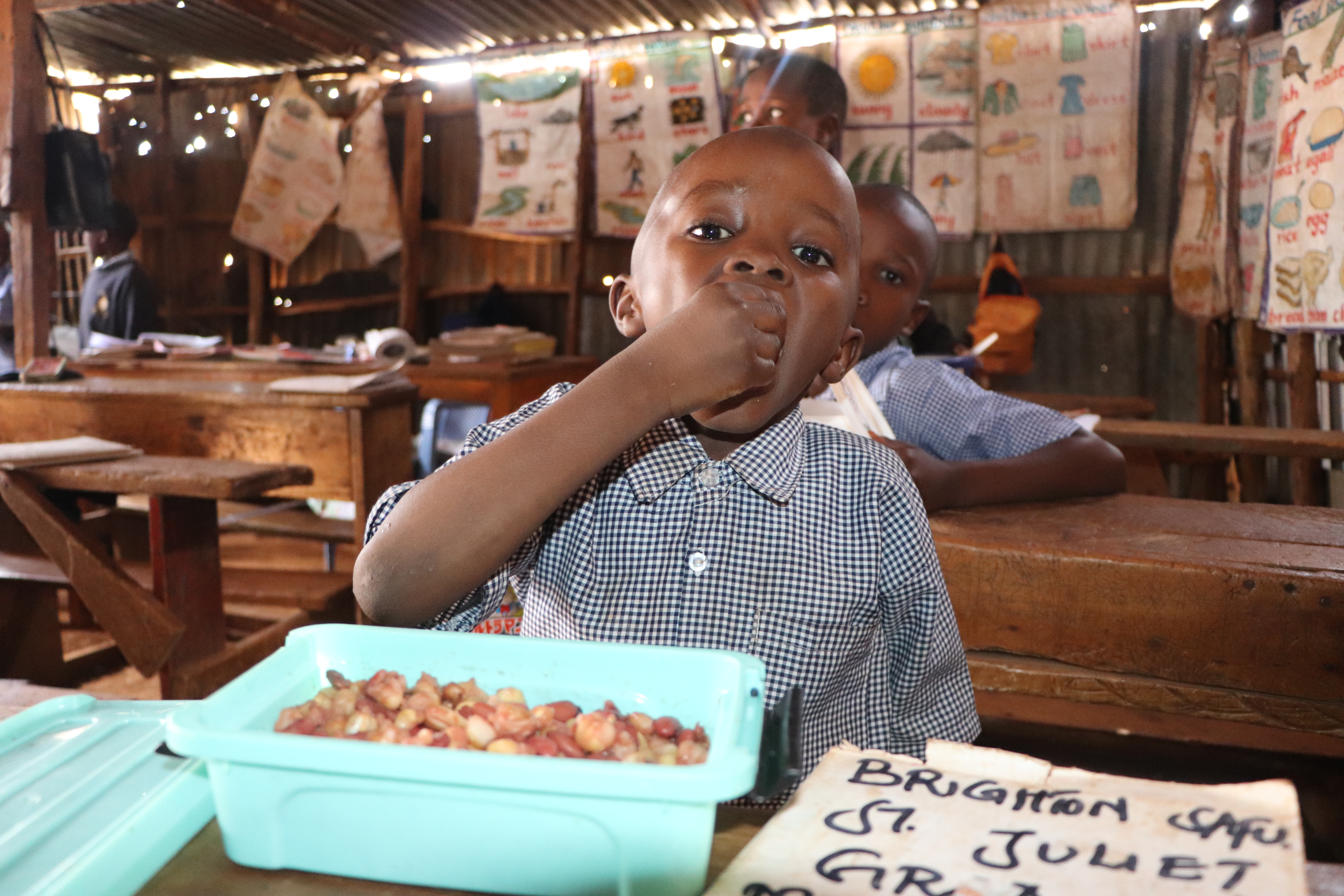 Ein Grundschulkind steckt sich Essen mit der Hand in den Mund. Es sitzt an einer Schulbank, auf der ein Kunststoffbehälter mit Essen steht.