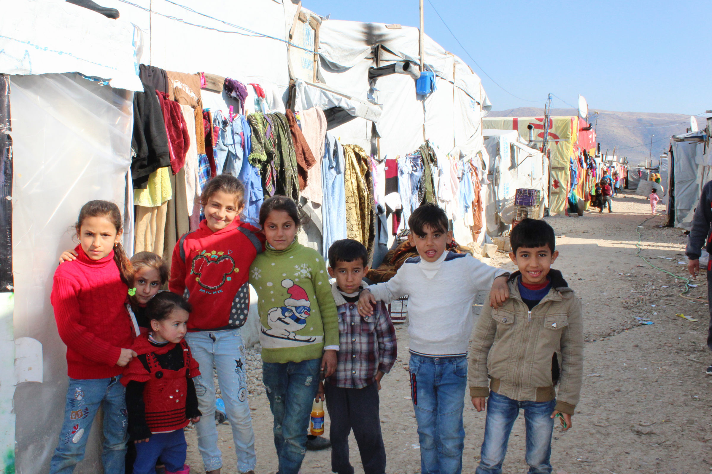 Eine Gruppe Kinder aufgereiht für das Foto, sie schauen in die Kamera. Im Hintergrund sieht man Zelte und provisorische Häuser