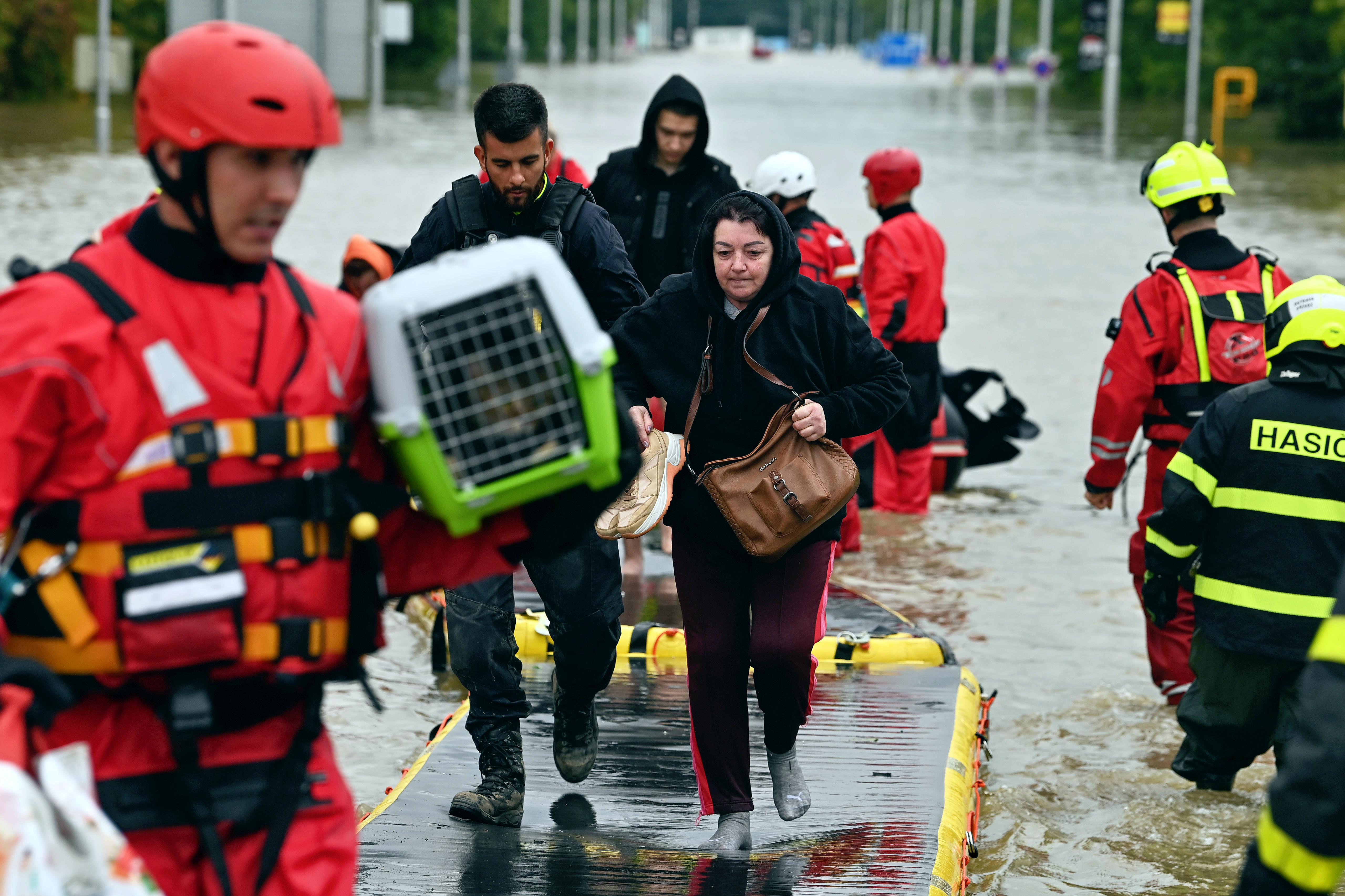 Vier Menschen in roter Uniform und mit Helmen begleiten eine kleine Gruppe Menschen, die durch Wasser watet. Ein Helfer trägt einen Korb für Haustiere.