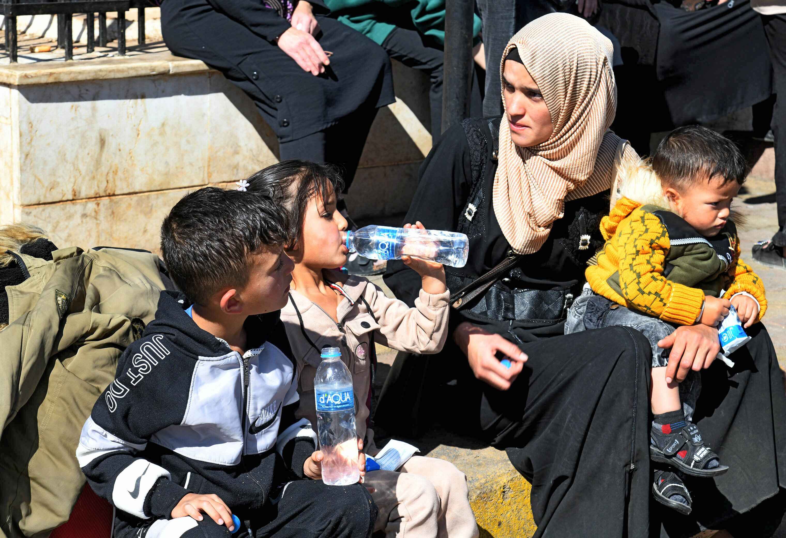 Frau und drei Kinder, eines davon ein Kleinkind, das auf dem Schoß der Frau sitzt. Die beiden anderen trinken Wasser aus Flaschen. Die Frau schaut zu den beiden Kindern.