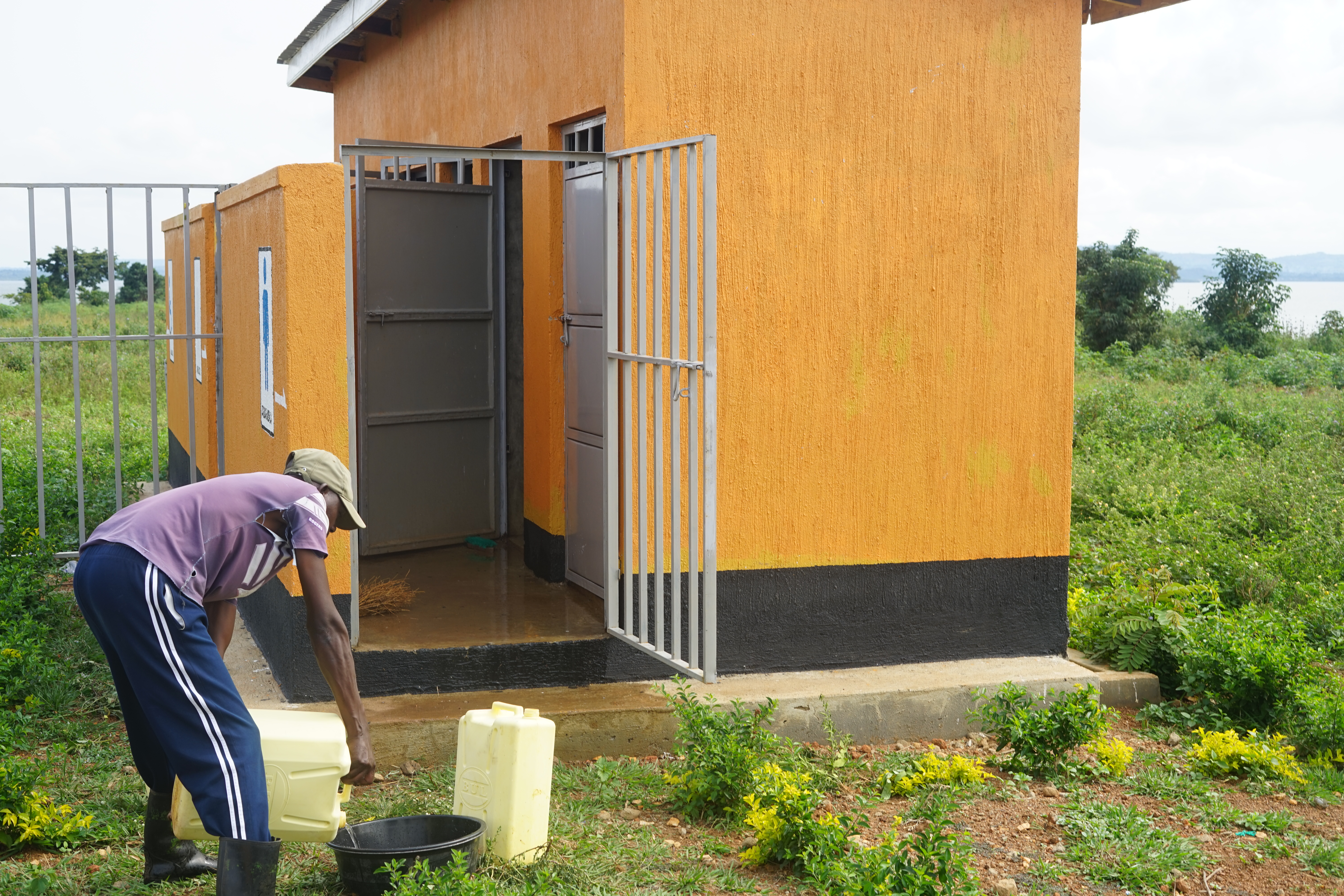 Mann kippt Wasser aus einem Kanister in eine Schüssel, er steht vor einem Sanitärgebäude