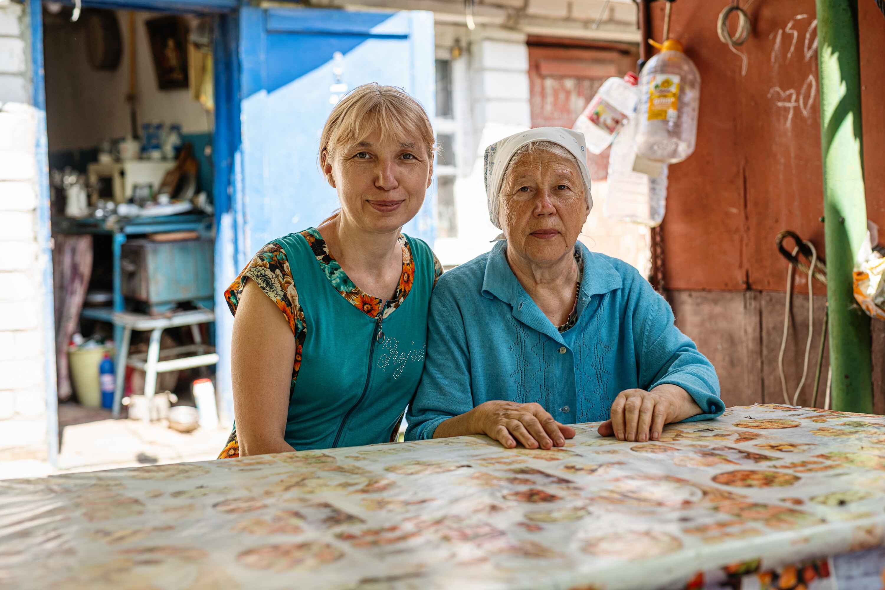Zwei Frauen, eine ältere und eine jüngere, sitzen an einem Tisch, im Hintergrund ist eine Art Geräteschuppen zu sehen.