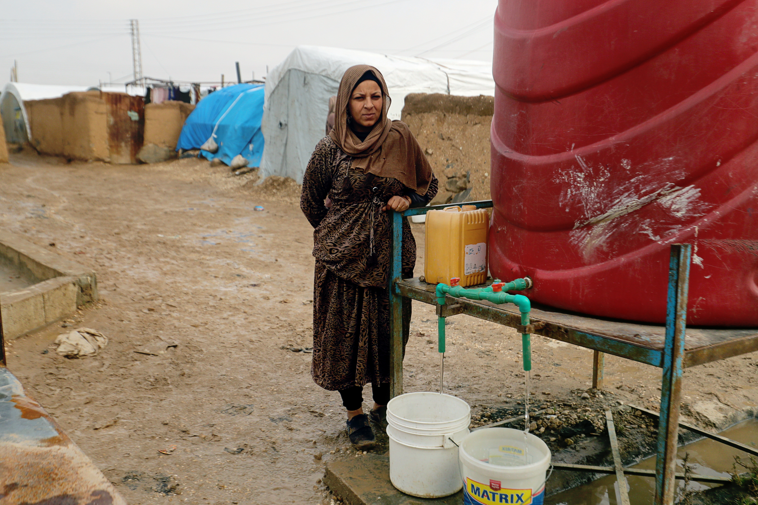 Frau steht an einem roten Wassertank in matschiger Umgebung und wartet darauf, dass ihr Eimer voll Wasser läuft.