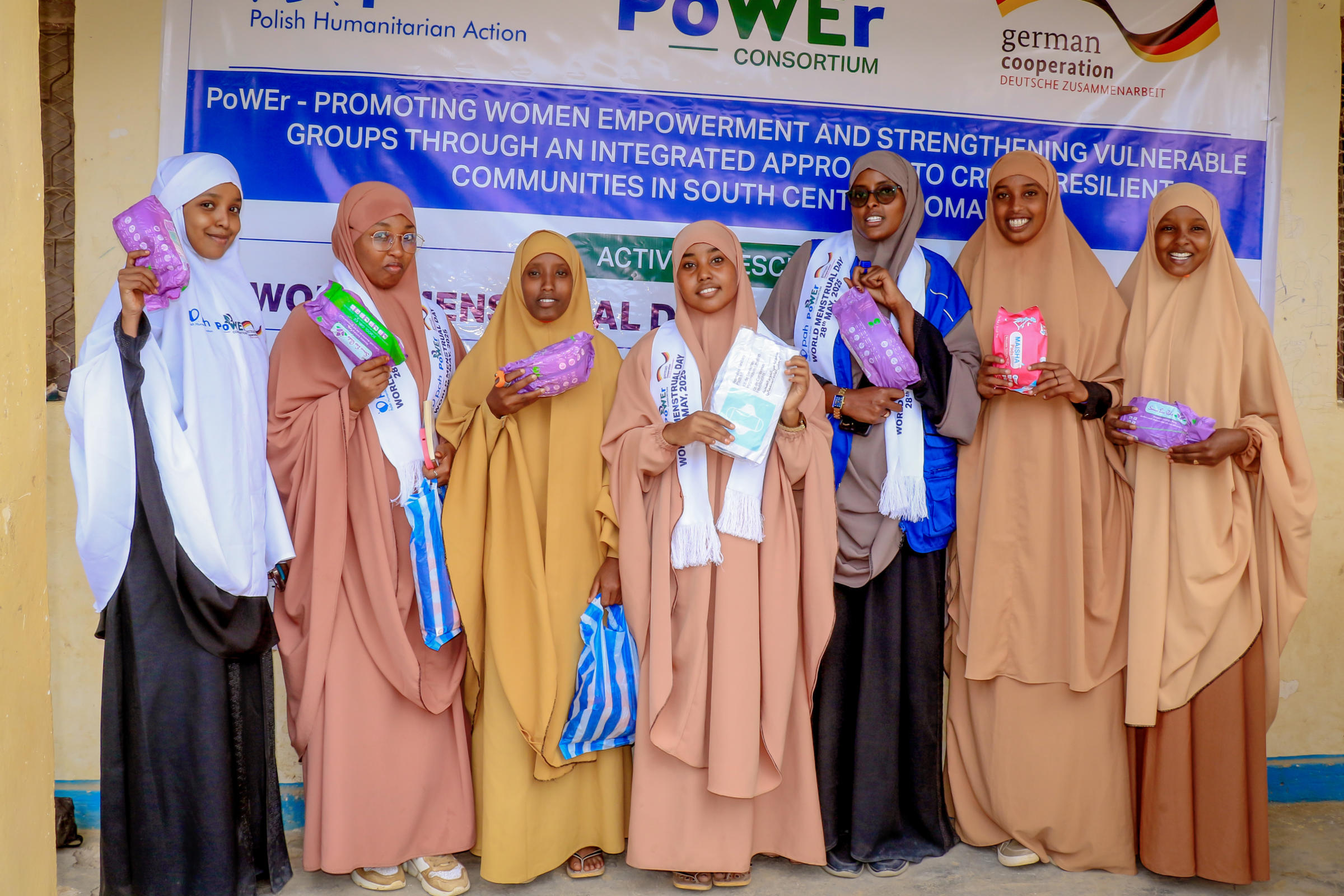 Sechs Frauen stehen vor einem Banner mit Schrift. Alle halten je ein Paket Damenbinden in der Hand und lächeln in die Kamera.