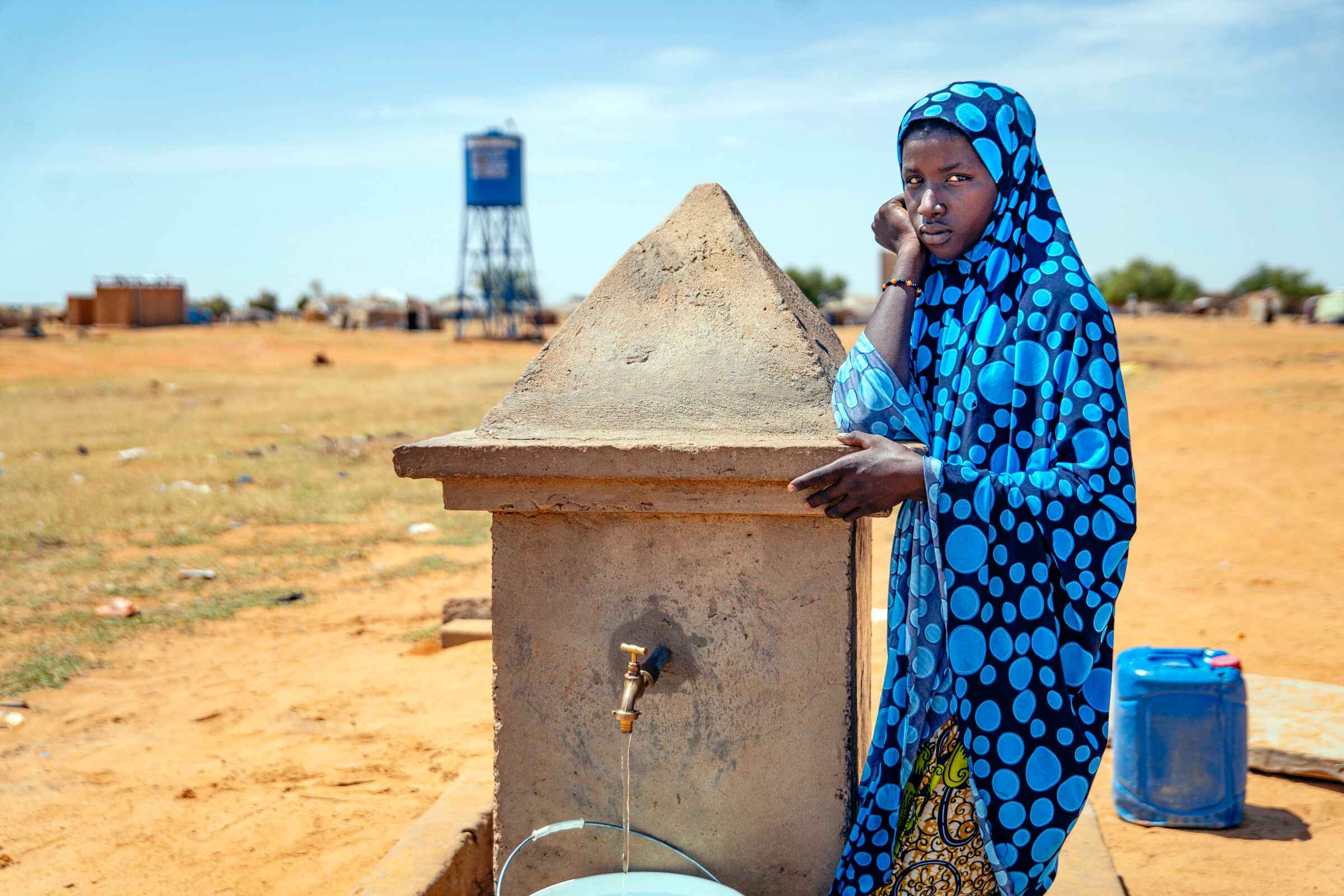 Frau lehnt an einem Bauwerk, das als Wasserausgabe dient und schaut in die Kamera.