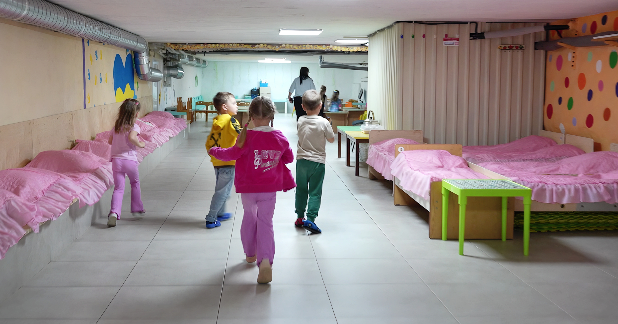 Innenraum des Kellers. Ein Kind sitzt auf einem der mehreren Betten mit rosa Bettdecken auf der linken Seite des Bildes, drei weitere Kinder laufen von der Kamera weg