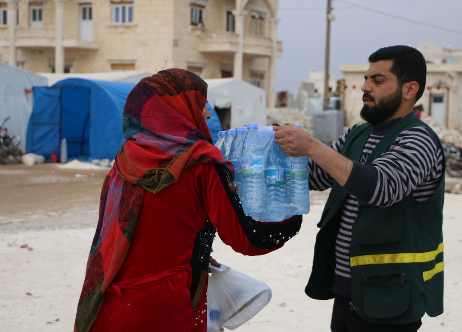 Mann (von schräg vorne) reicht einer Frau (von schräg hinten) ein Gebinde von Kunststoffflaschen mit Wasser
