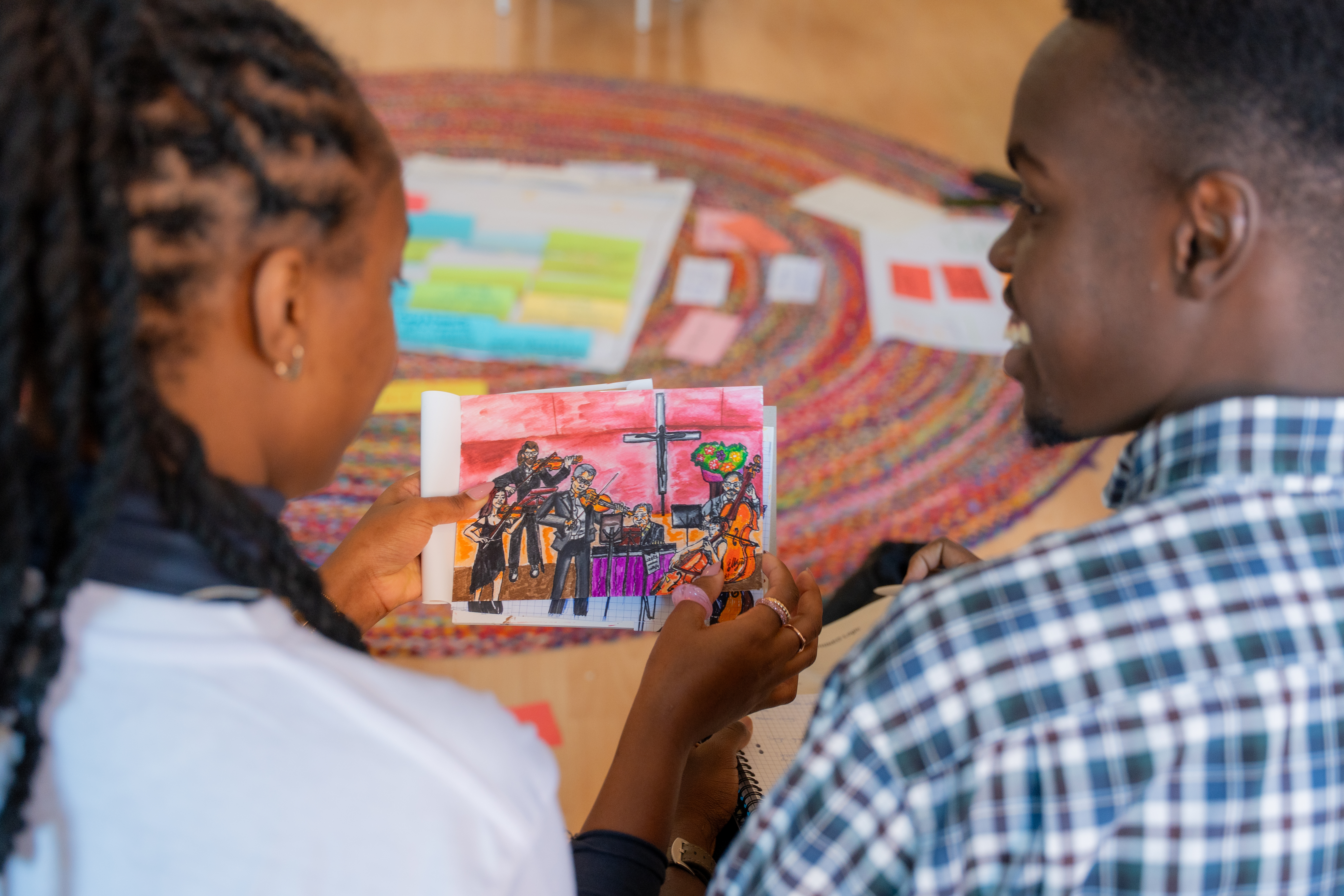 Zwei Persons of Colour sitzen nebeneinander,eine von ihnen hält ein gezeichnetes Bild von weißen Musiker*innen in der Hand. Die Aufnahme zeigt sie von hinten im Profil, im Hintergrund ist zu sehen, dass Papier am Boden liegt.