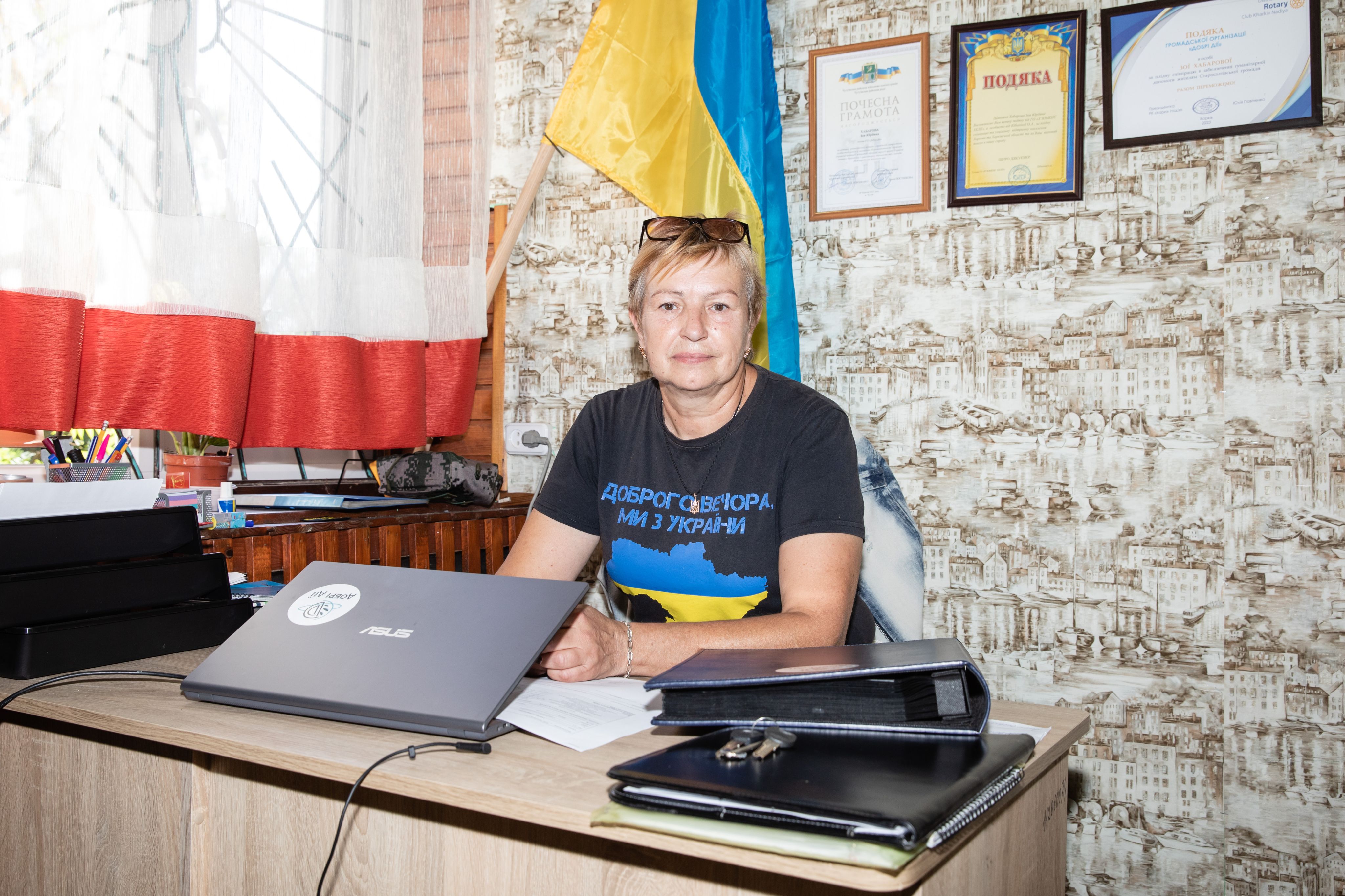 Im Bild ist Zoia Khabarova zu sehen, die am Schreibtisch ihres Büros sitzt. Sie trägt ein Shirt, auf dem die Länderumrisse der Ukraine in den Flaggenfarben abgebildet sind.