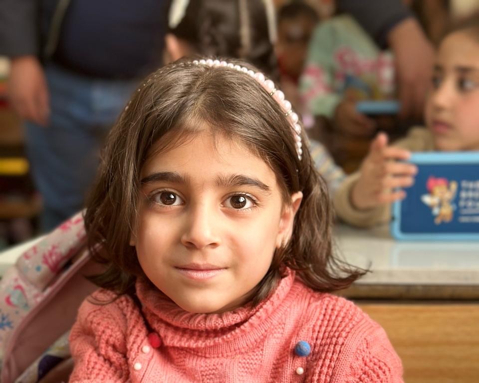 Ein ungefähr 6 Jahre altes Mädchen im Klassenzimmer, freundlicher Blick, im Hintergrund andere Schüler*innen