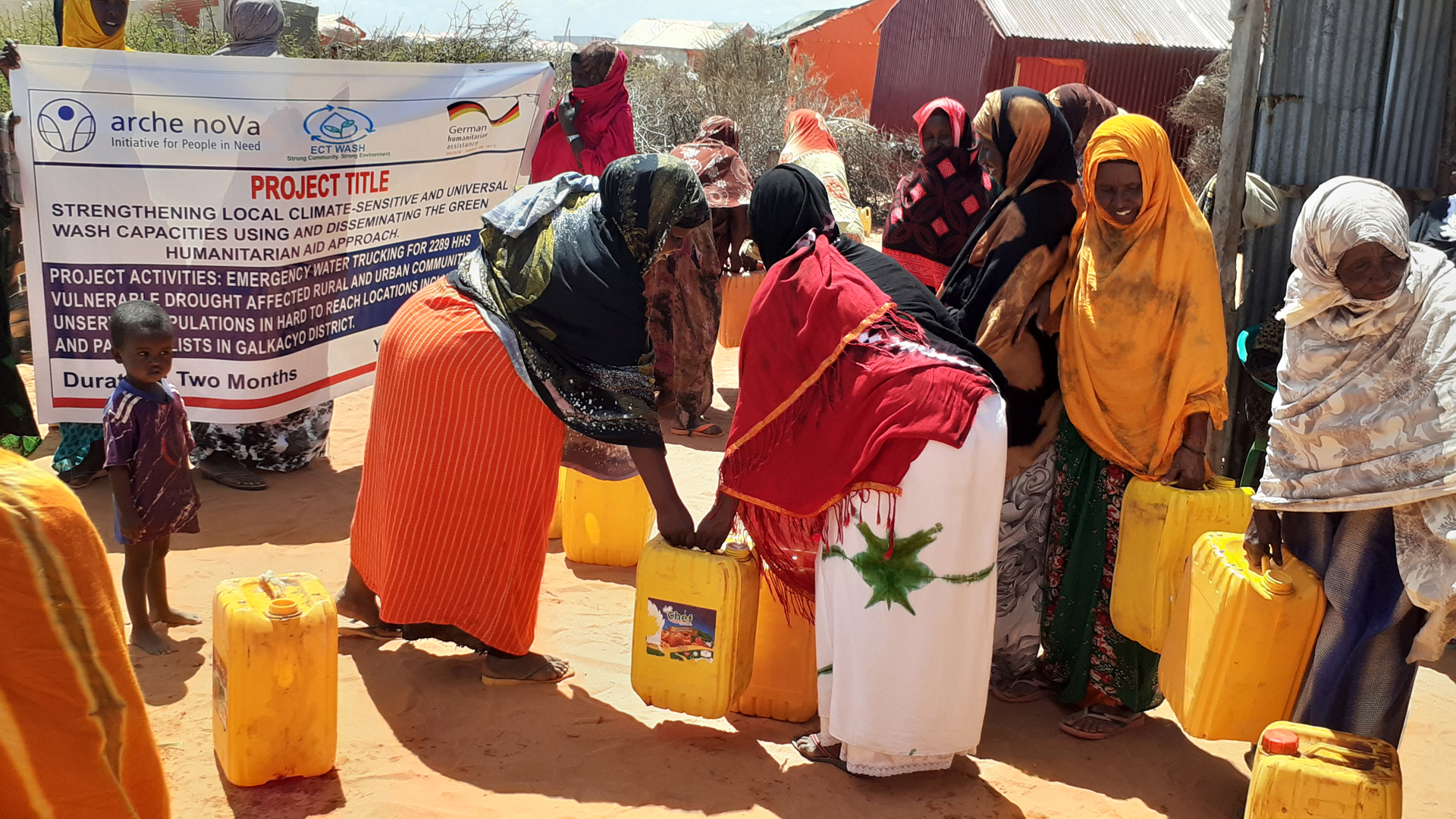 Eine Gruppe Frauen im Freien, die gelbe Wasserkanister aufheben oder tragen. Im Hintergrund halten zwei Personen ein Banner, auf dem der Projekttitel des Global Programms zu lesen ist. 