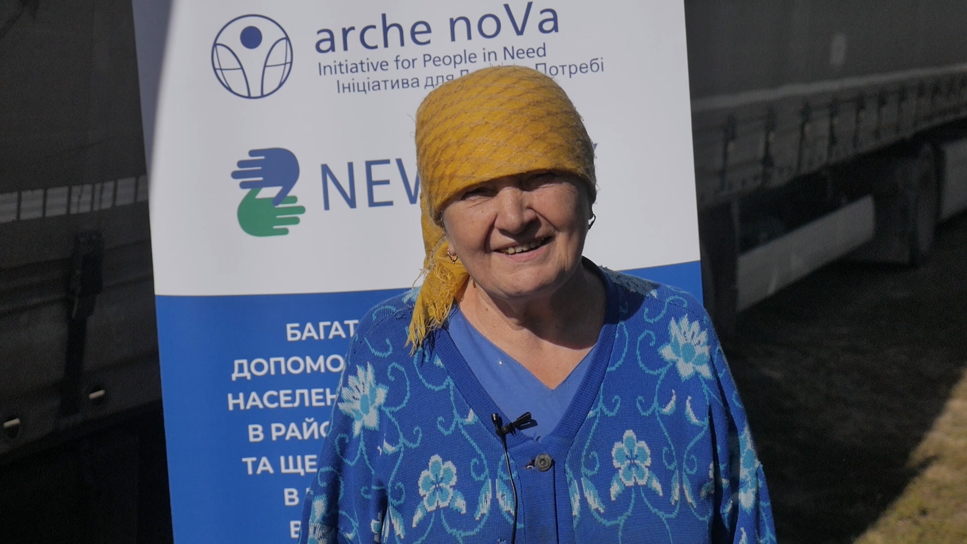•	Portrait einer älteren Frau mit gelbem Kopftuch im Hintergrund ein Plakat von arche noVa 