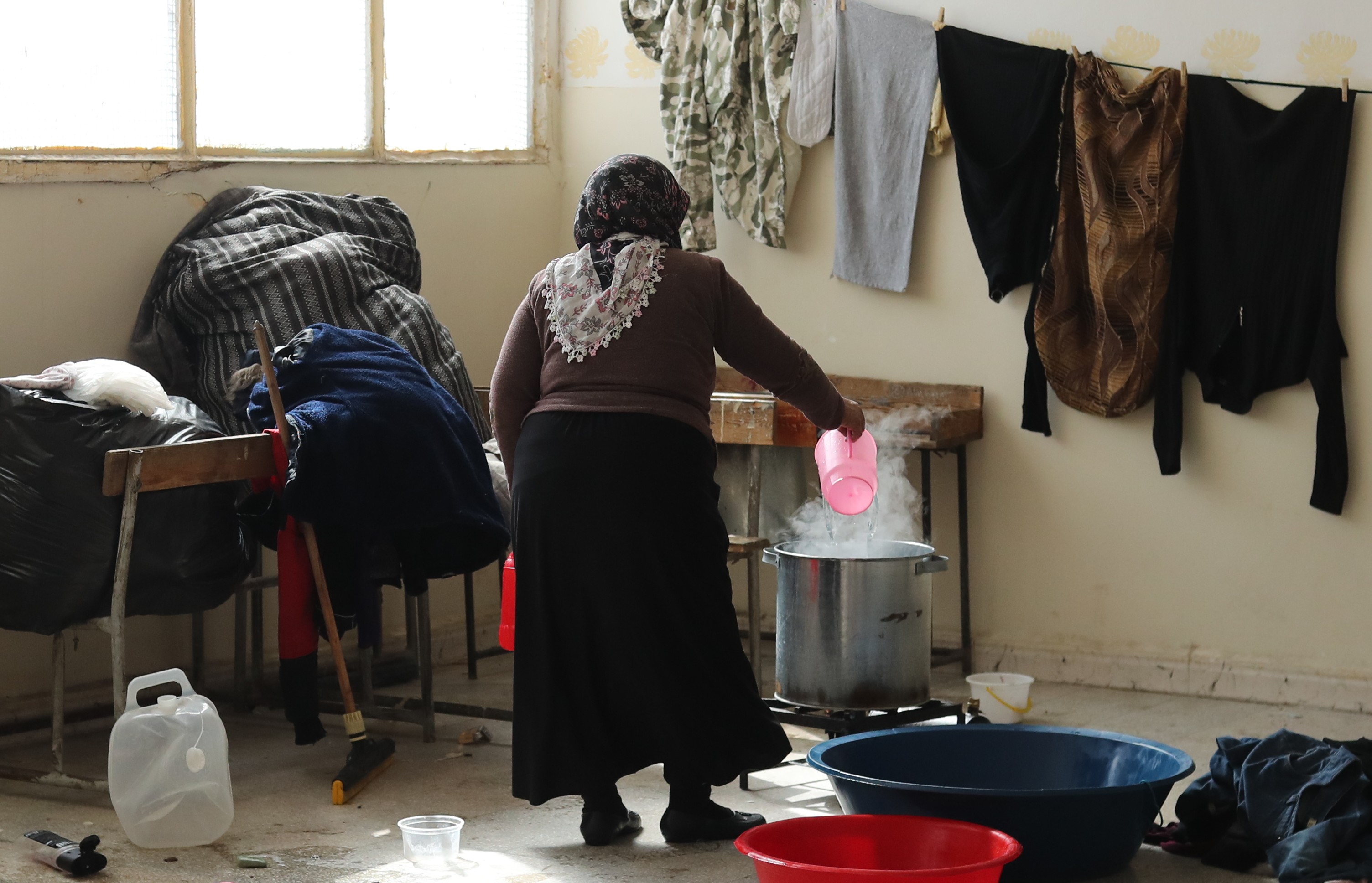 eine Frau in einer Zimmerecke gießt Wasser in einen großen Topf, der auf einer Kochplatte am Boden steht