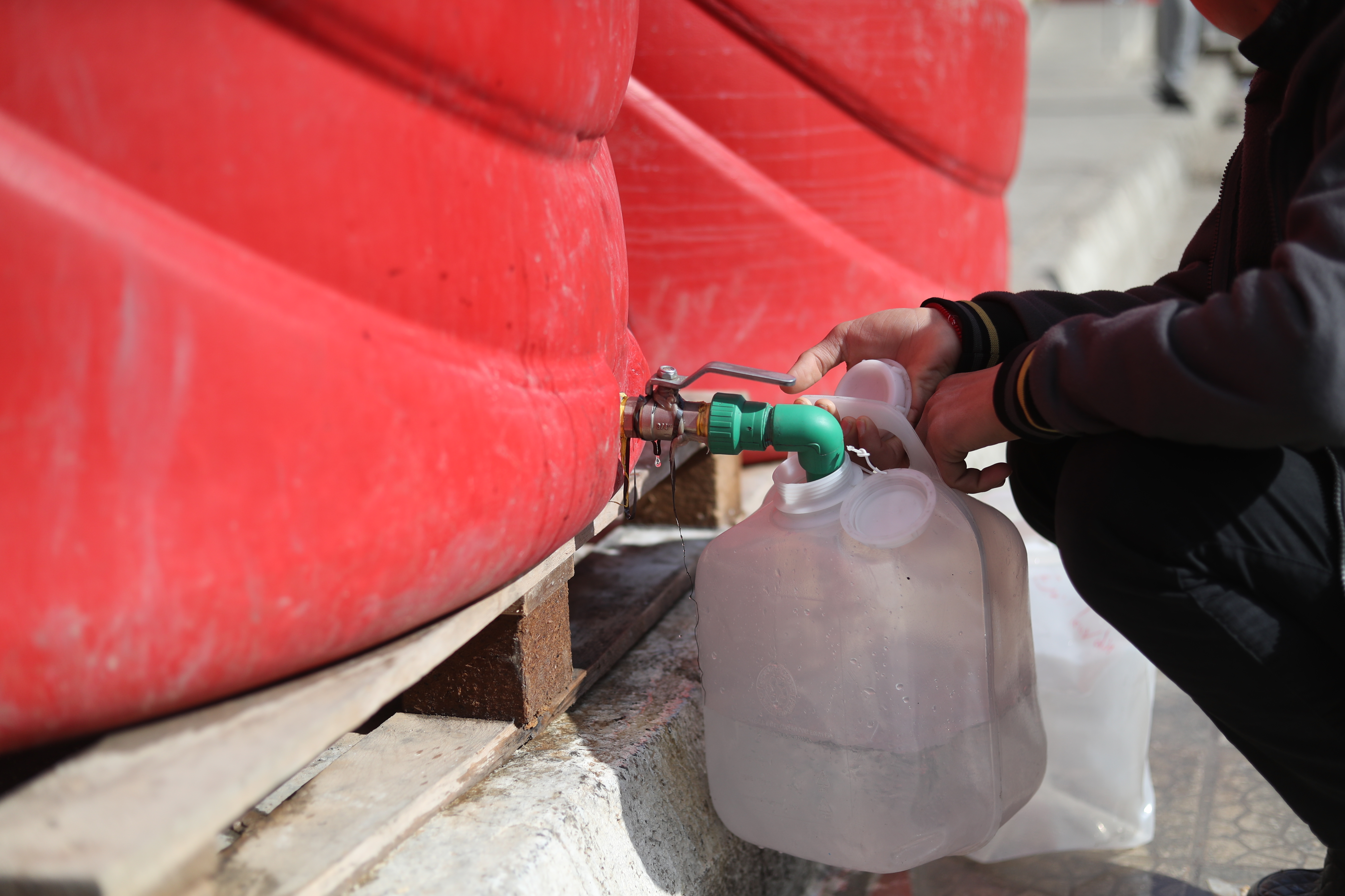 Wasserhahn am unteren Ende eines großen roten Wassertanks, gerade fließt Wasser in einen Kanister