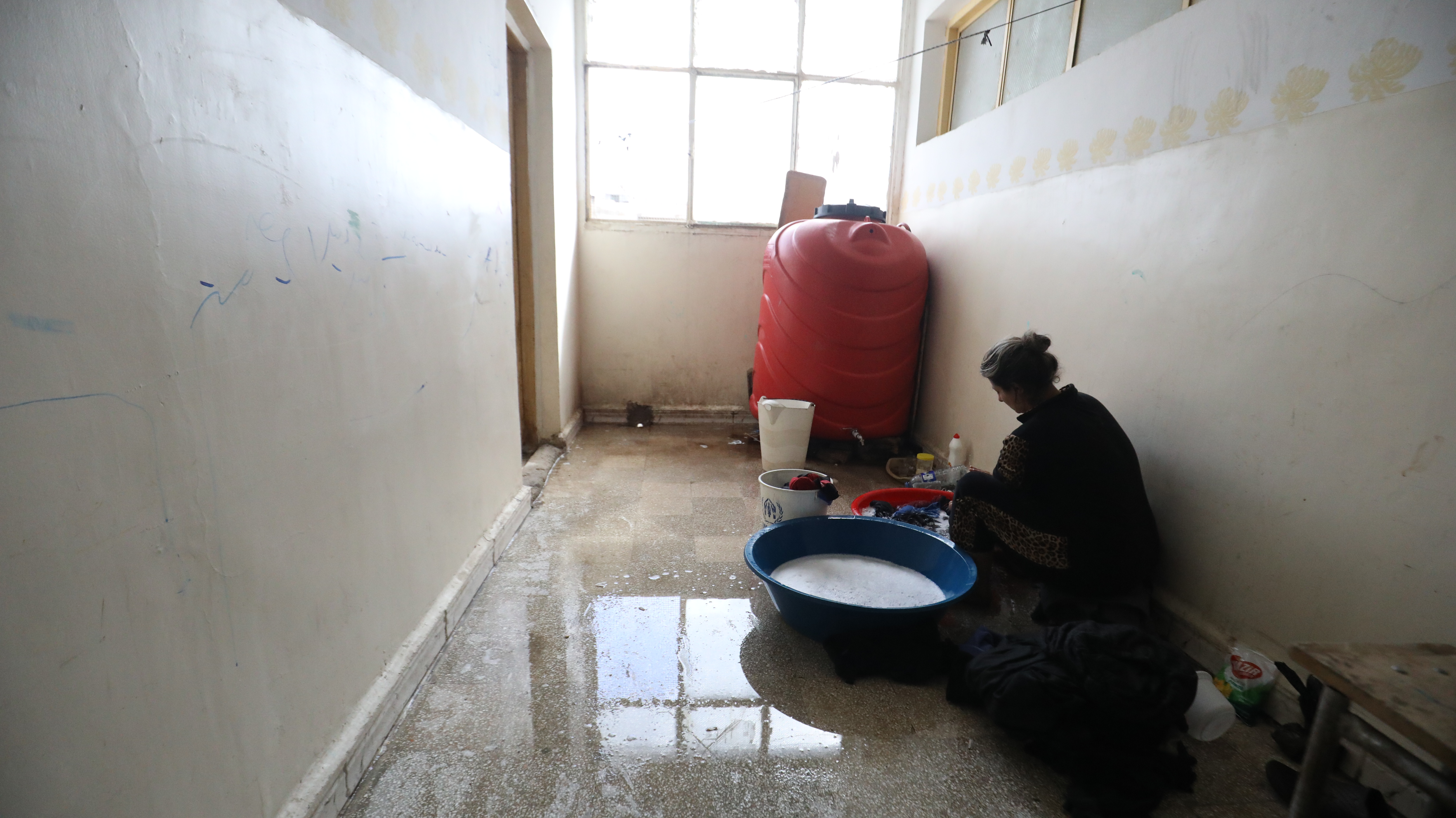 Am Ende eines Flurs steht ein roter Wassertank, davor hockt eine Frau vor einer Waschschüssel