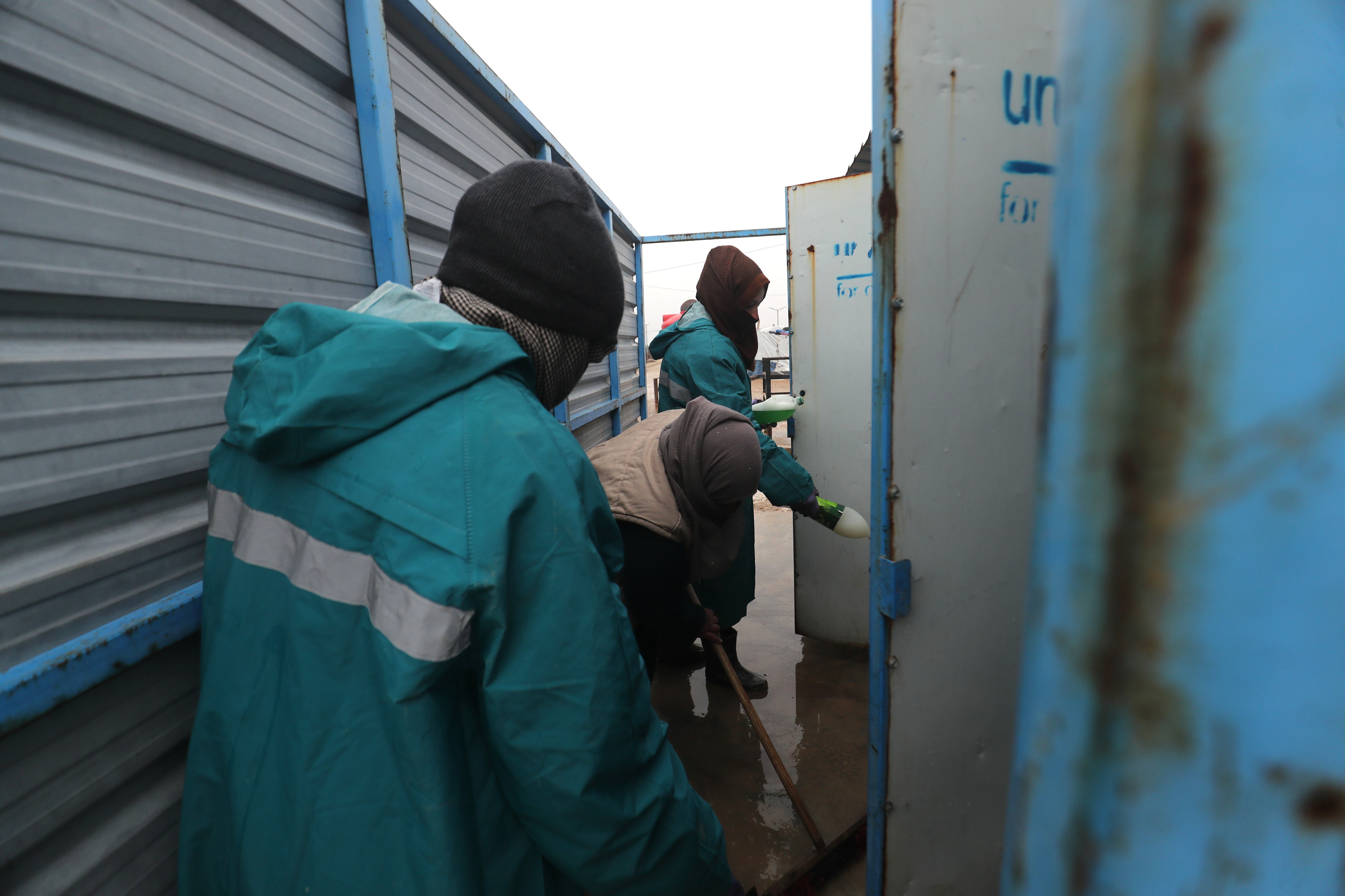 Zwischen zwei Wellblechgebäuden stehen drei Menschen in Regenjacken