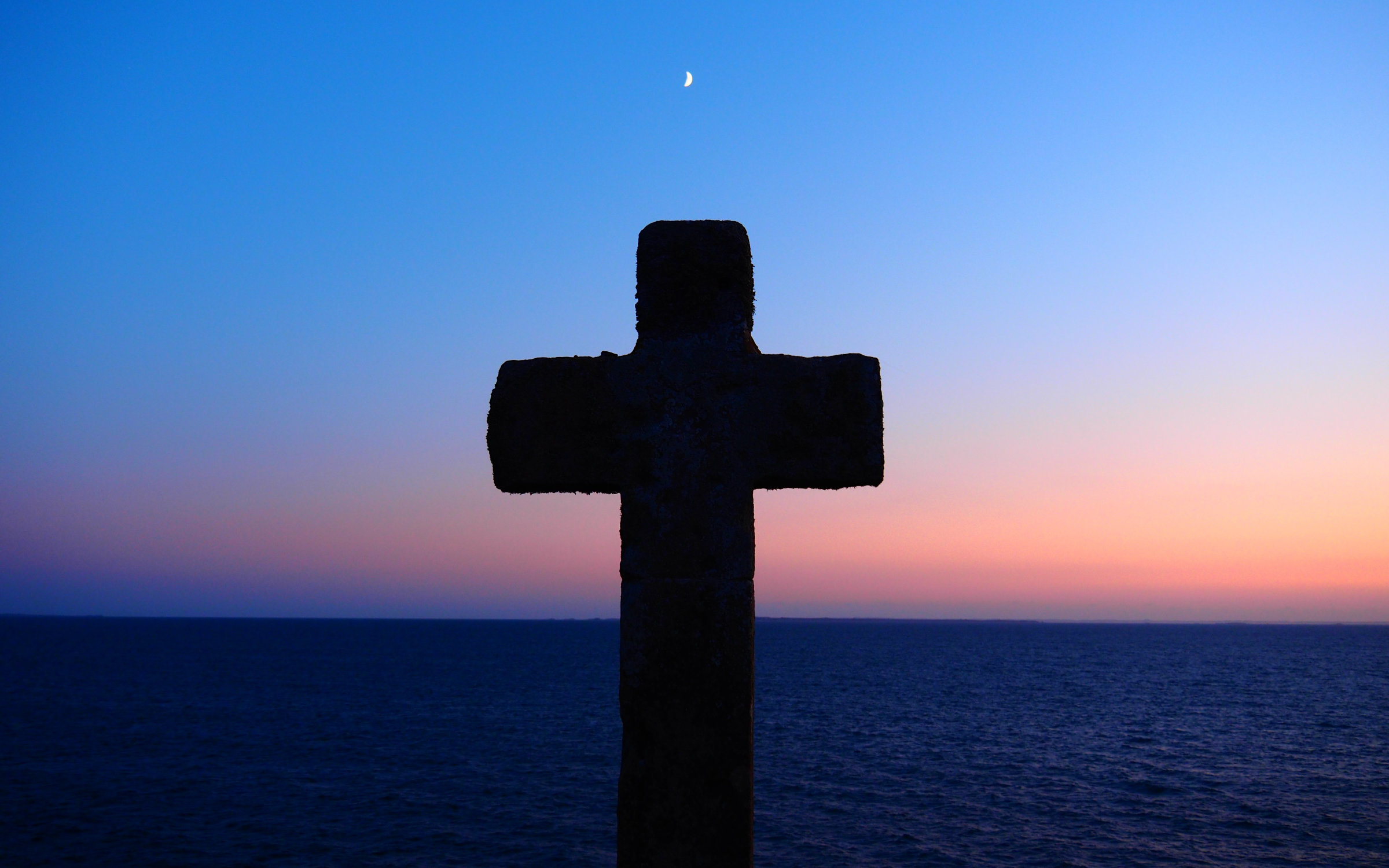 Szenerie eines Sonnenuntgergangs, zentral im Vordergrund ein massives Kreuz, darüber eine Mondichel